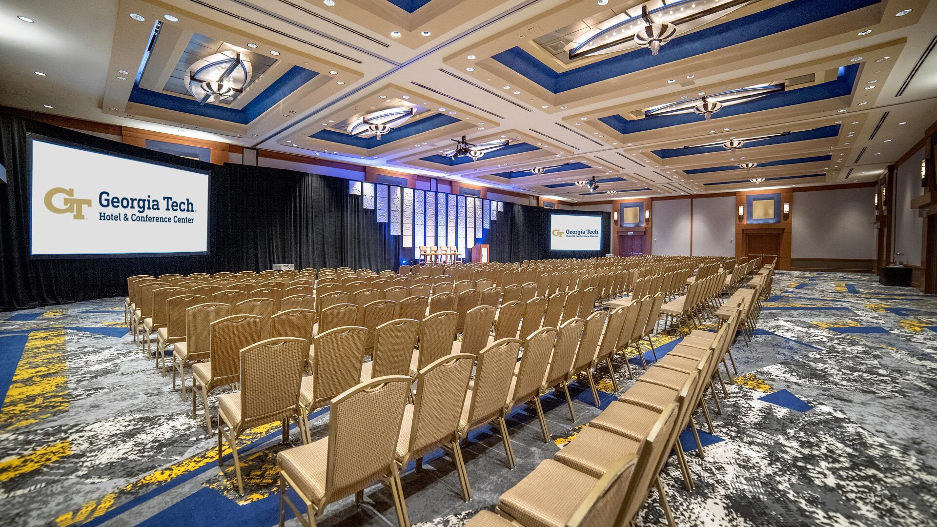 A conference room with rows of chairs, a large screen displaying 'Georgia Tech Hotel & Conference Center', and a patterned carpet.