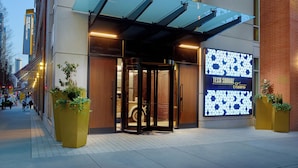 Entrada d'un hotel modern amb façana de vidre, un panell informatiu digital i una zona d'accés coberta.