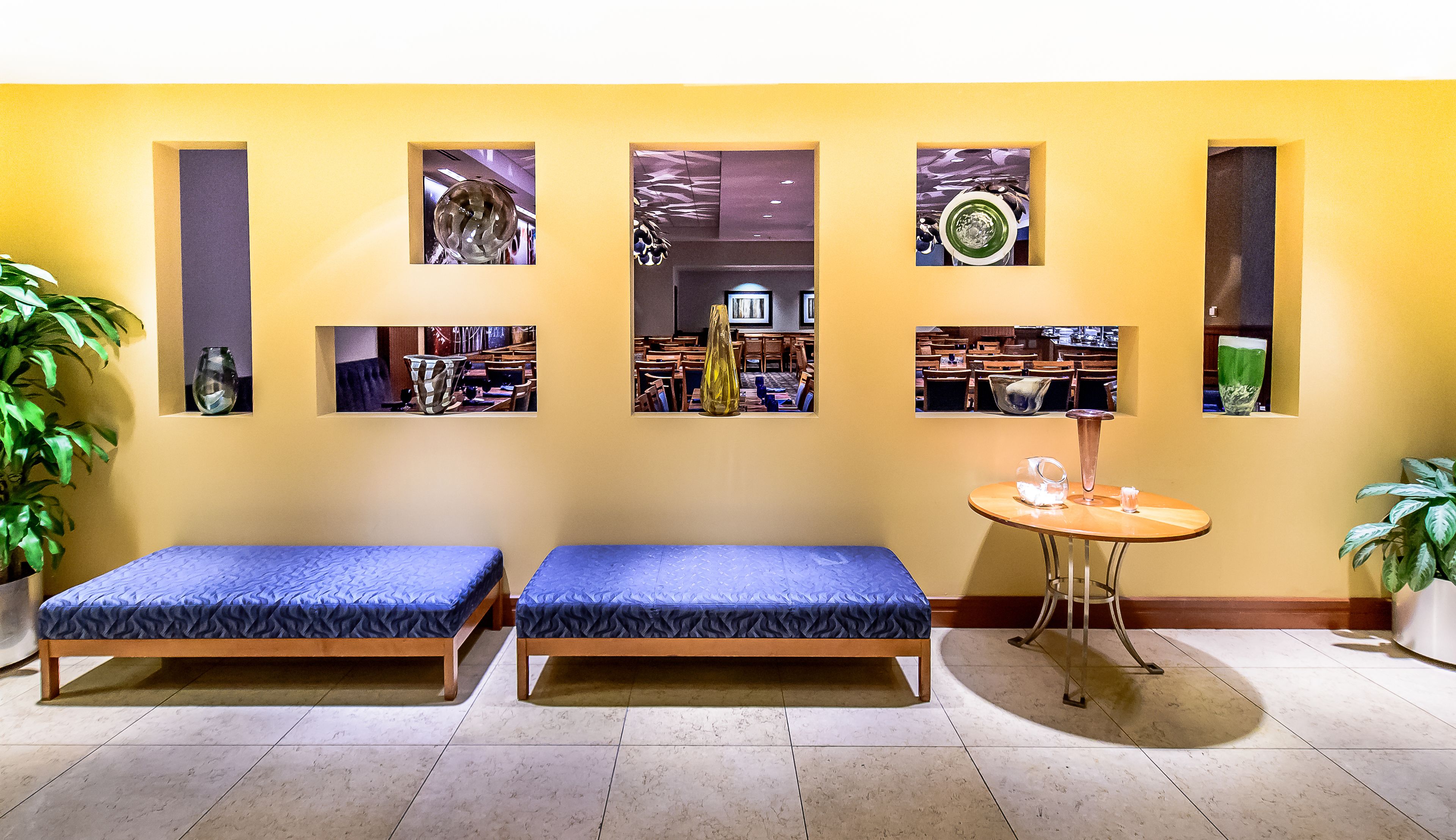 Une pièce avec deux lits recouverts de coussins bleus, une petite table ronde ornée d’un vase et d’une bougie, et trois tableaux encadrés accrochés au mur.
