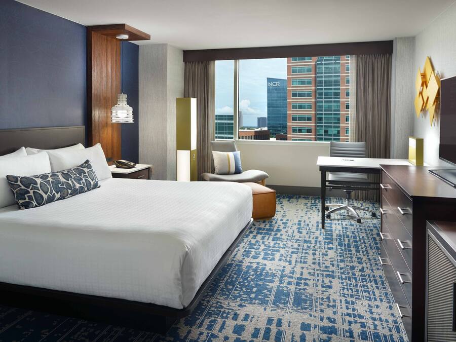 A hotel room with a large bed, a desk, a chair, a television, and a view of the cityscape.