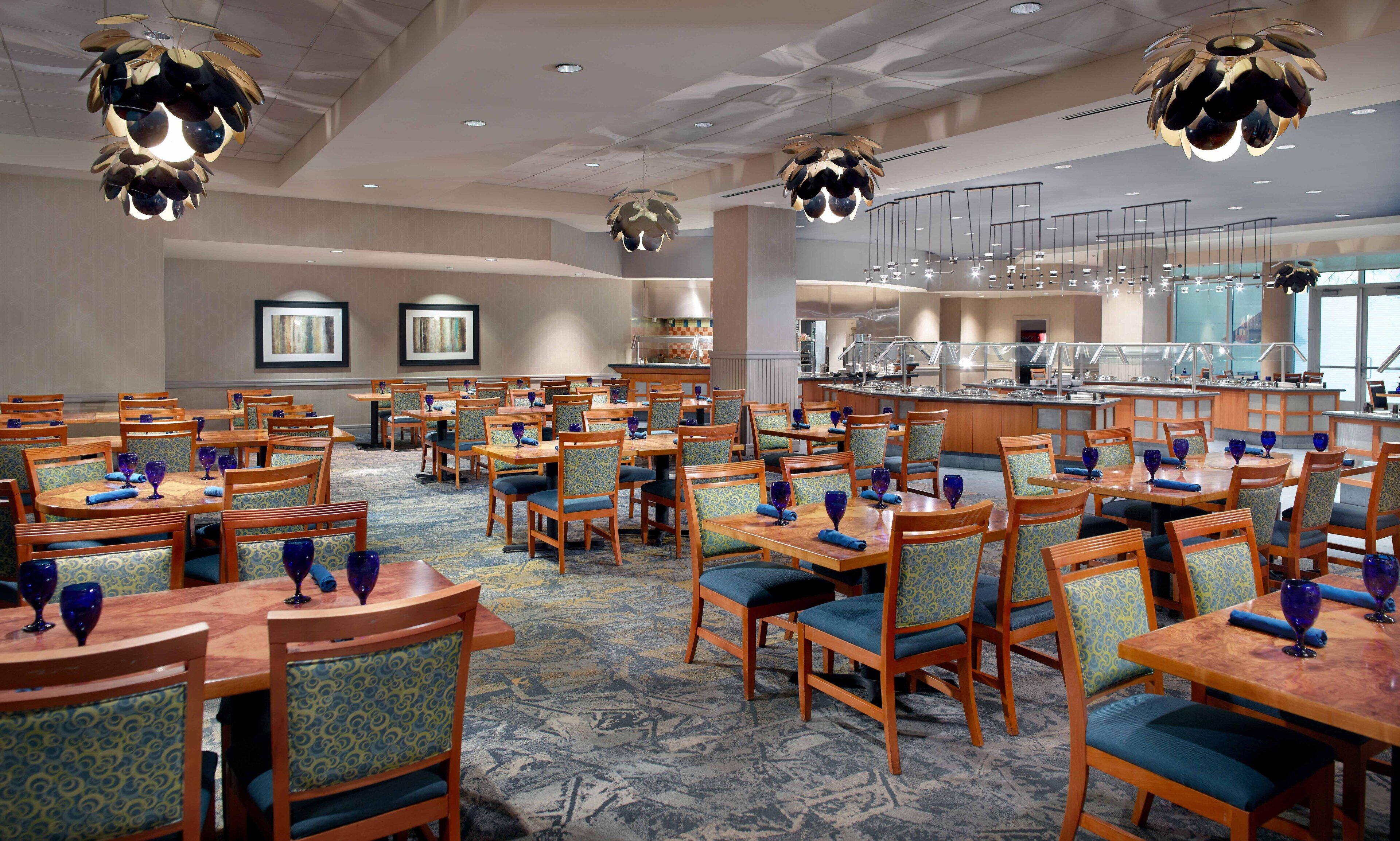 Zona de comedor de hotel con varias mesas y sillas, una barra de buffet e iluminación colgante moderna.