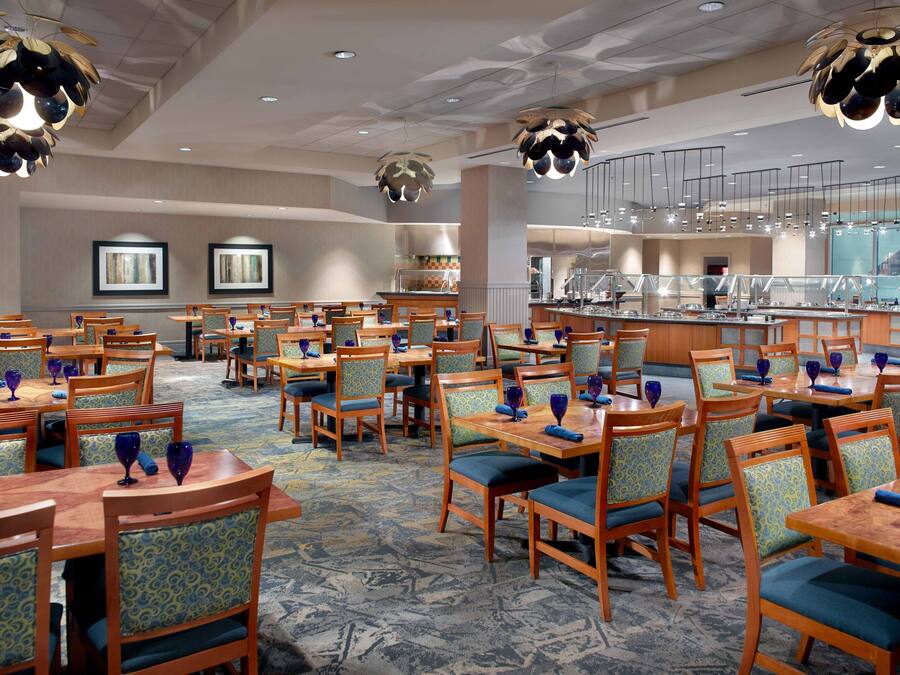 Zona de comedor de hotel con varias mesas y sillas, una barra de buffet e iluminación colgante moderna.