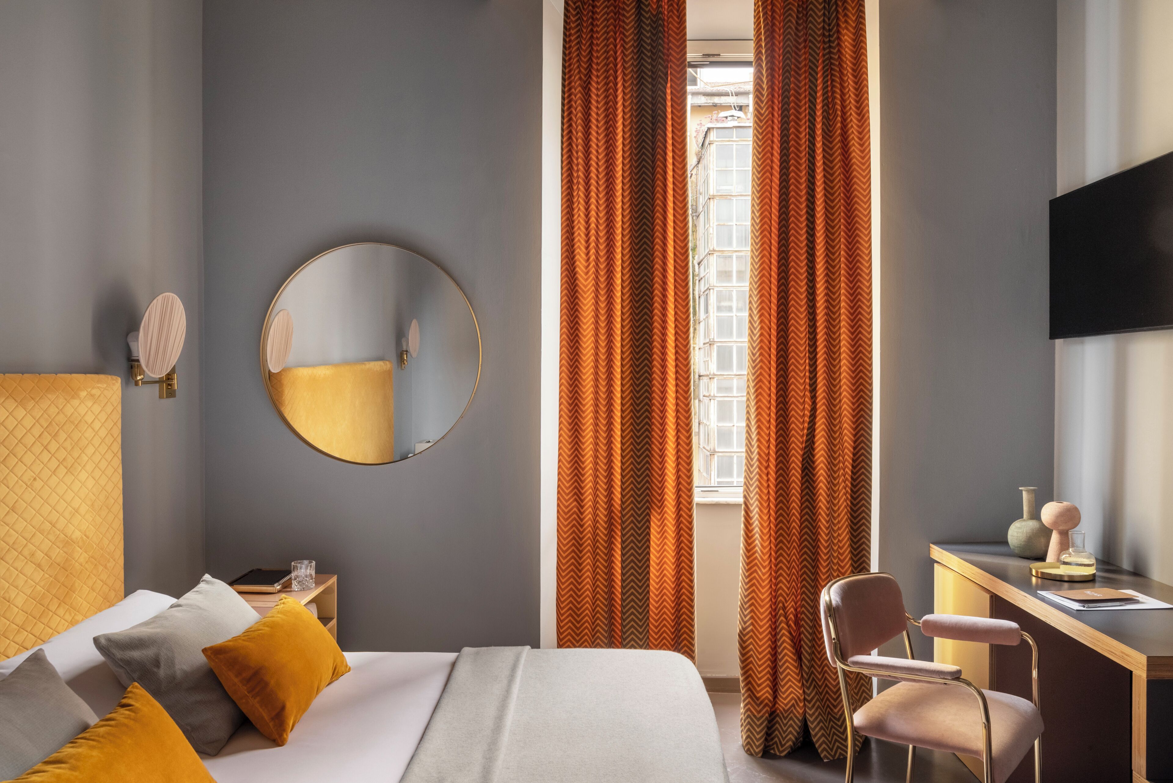 Habitación de hotel con cama, escritorio, silla, espejo redondo y ventana con cortinas.