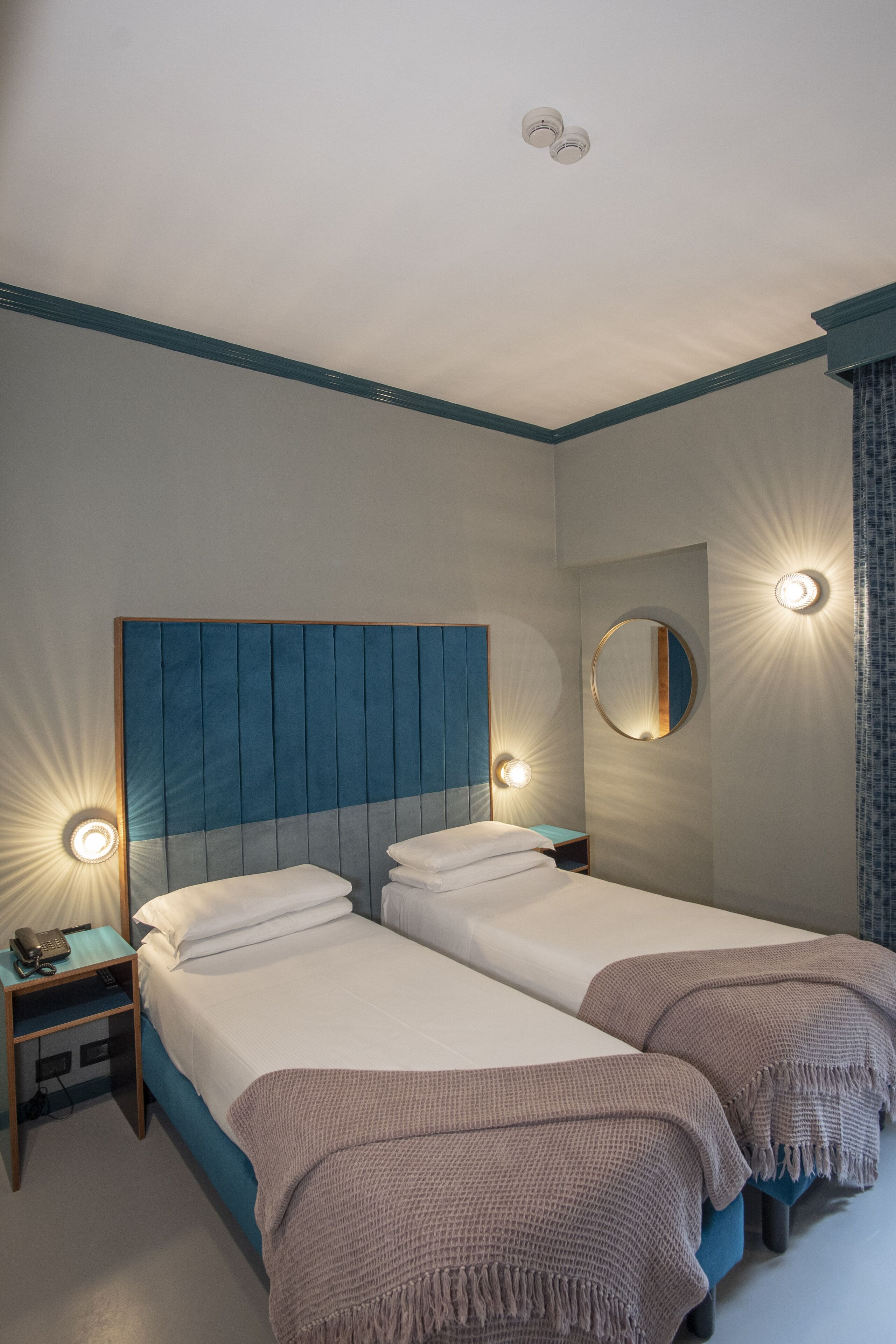 A hotel room with two beds, a round mirror, and wall sconces.
