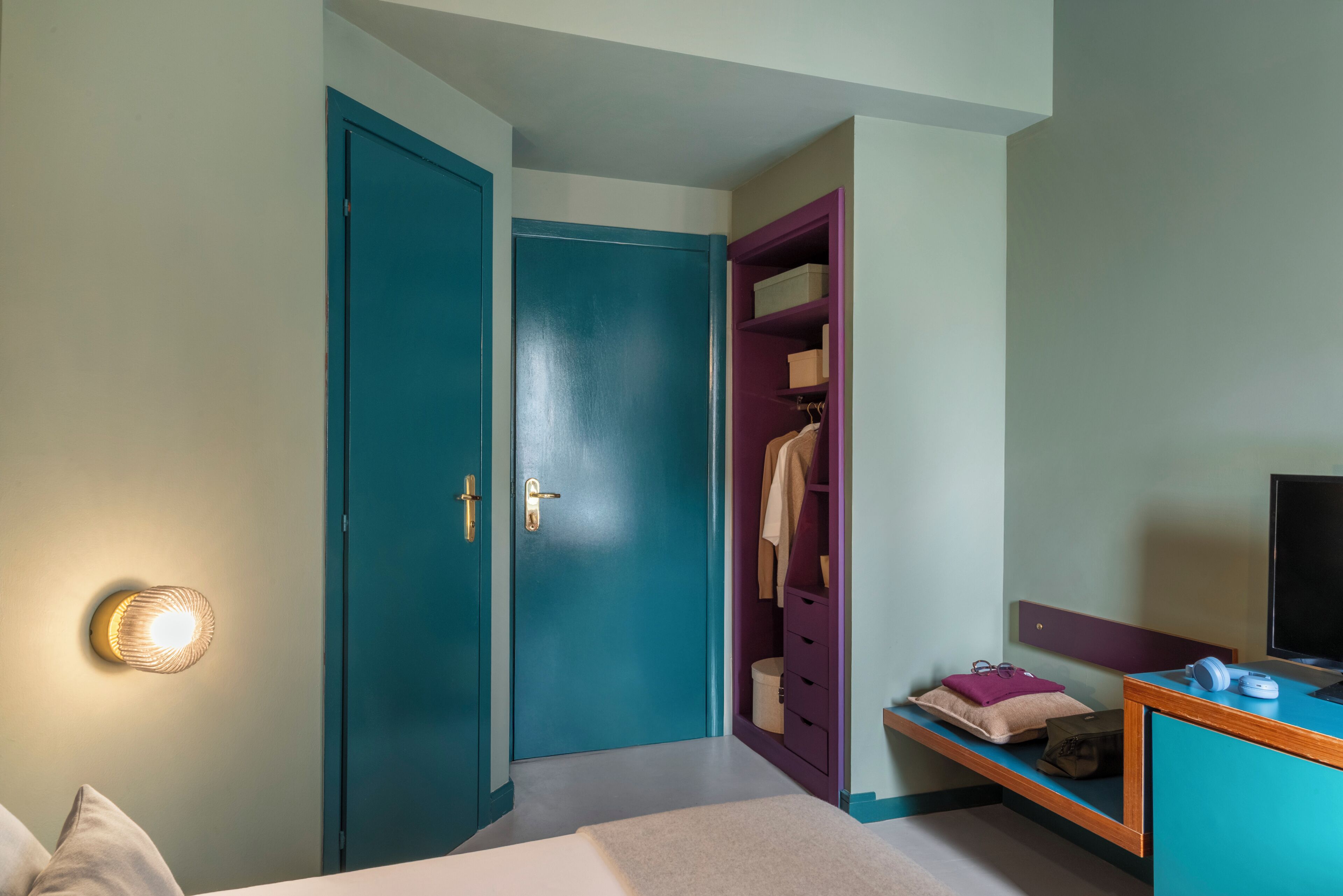 A bedroom with a teal door, a closet with purple interior, a bedside table with headphones, and a TV on the right.