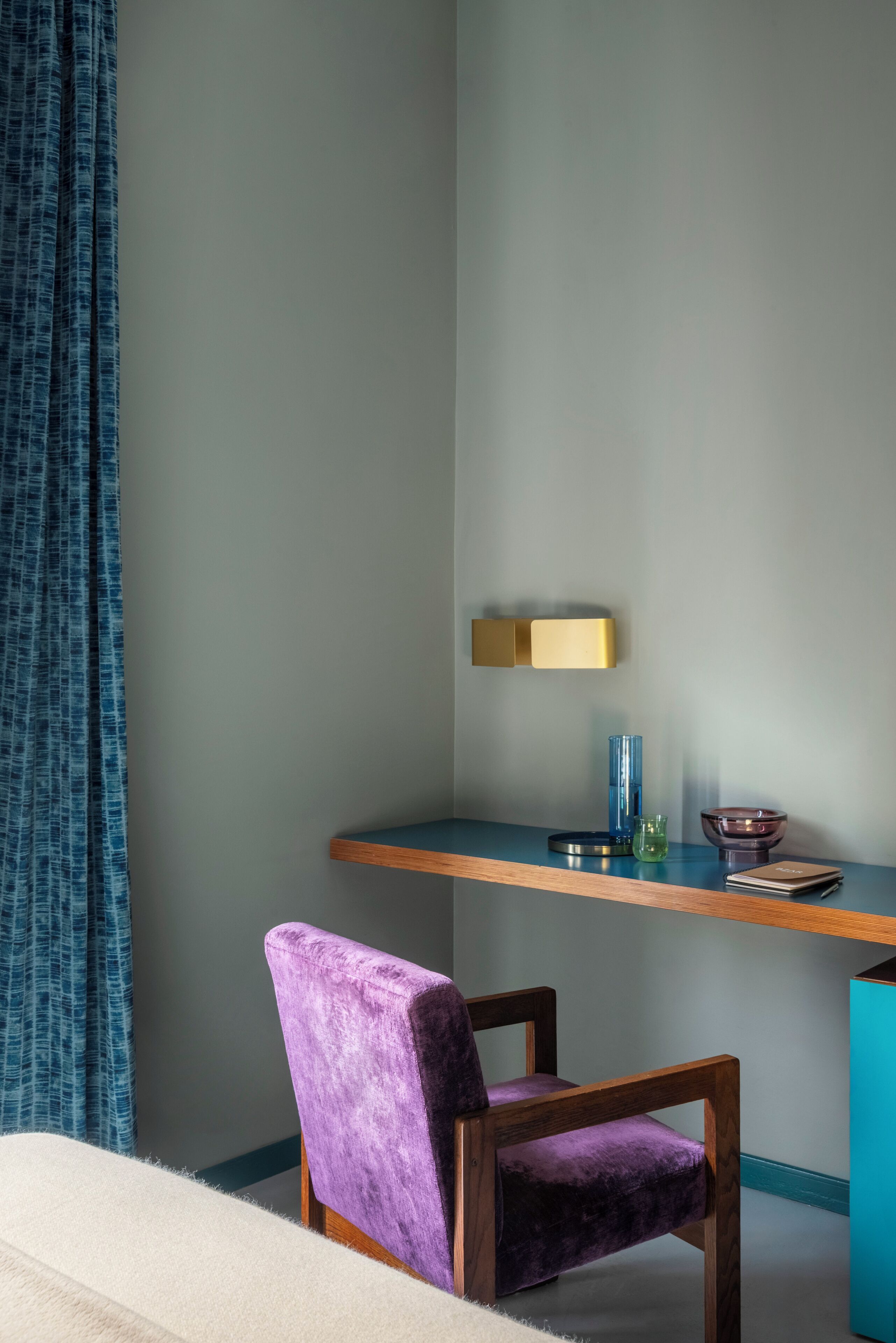 Une pièce d’angle avec un fauteuil violet, un bureau en bois, un vase bleu et un luminaire fixé au mur.