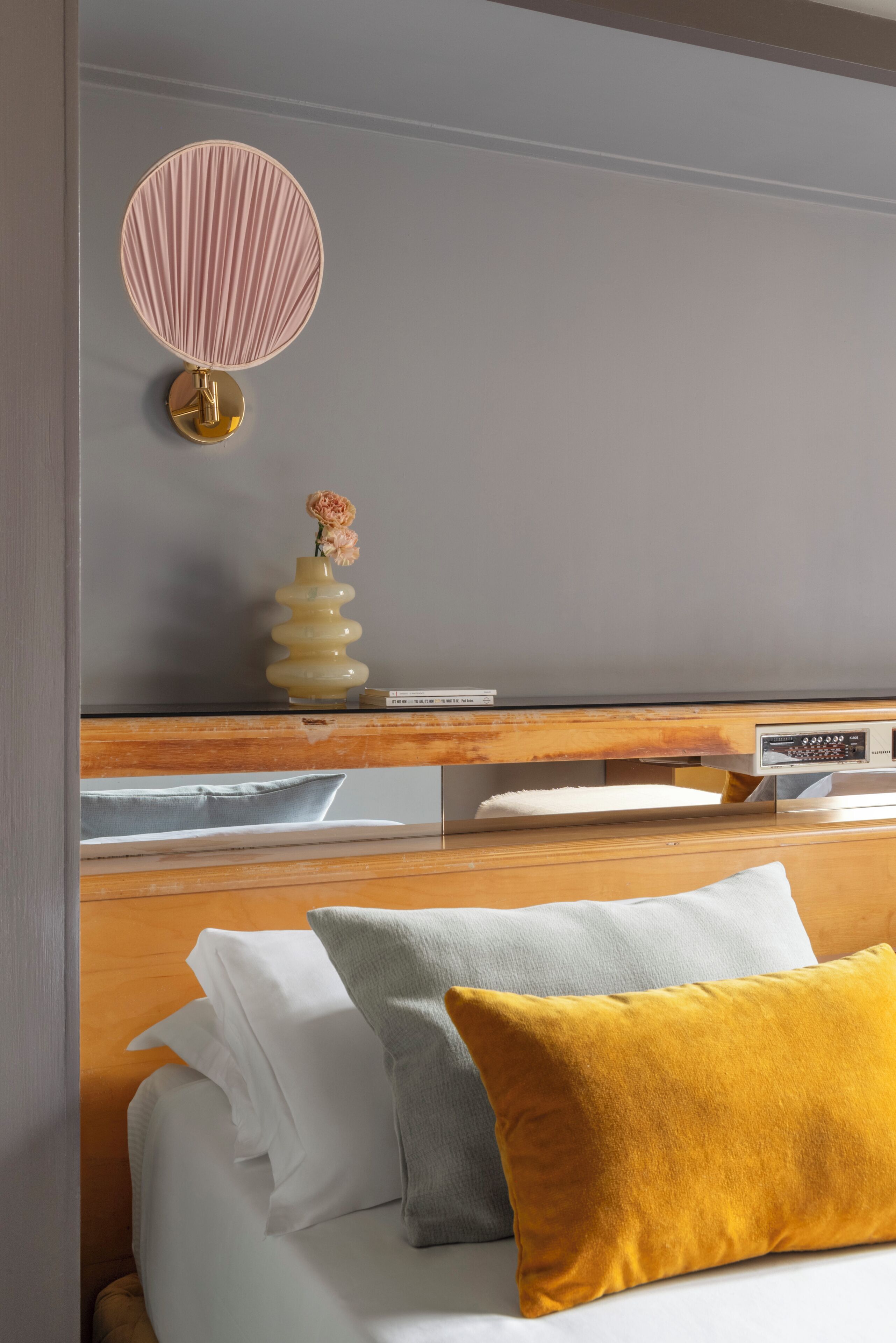 A hotel room with a bed, pillows, a bedside table with a lamp, a vase with flowers, and a wall-mounted clock.