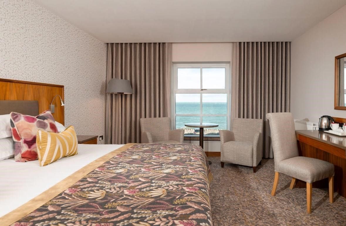 A hotel room with a bed, two chairs, a desk, a lamp, a view of the sea, and a window with curtains.