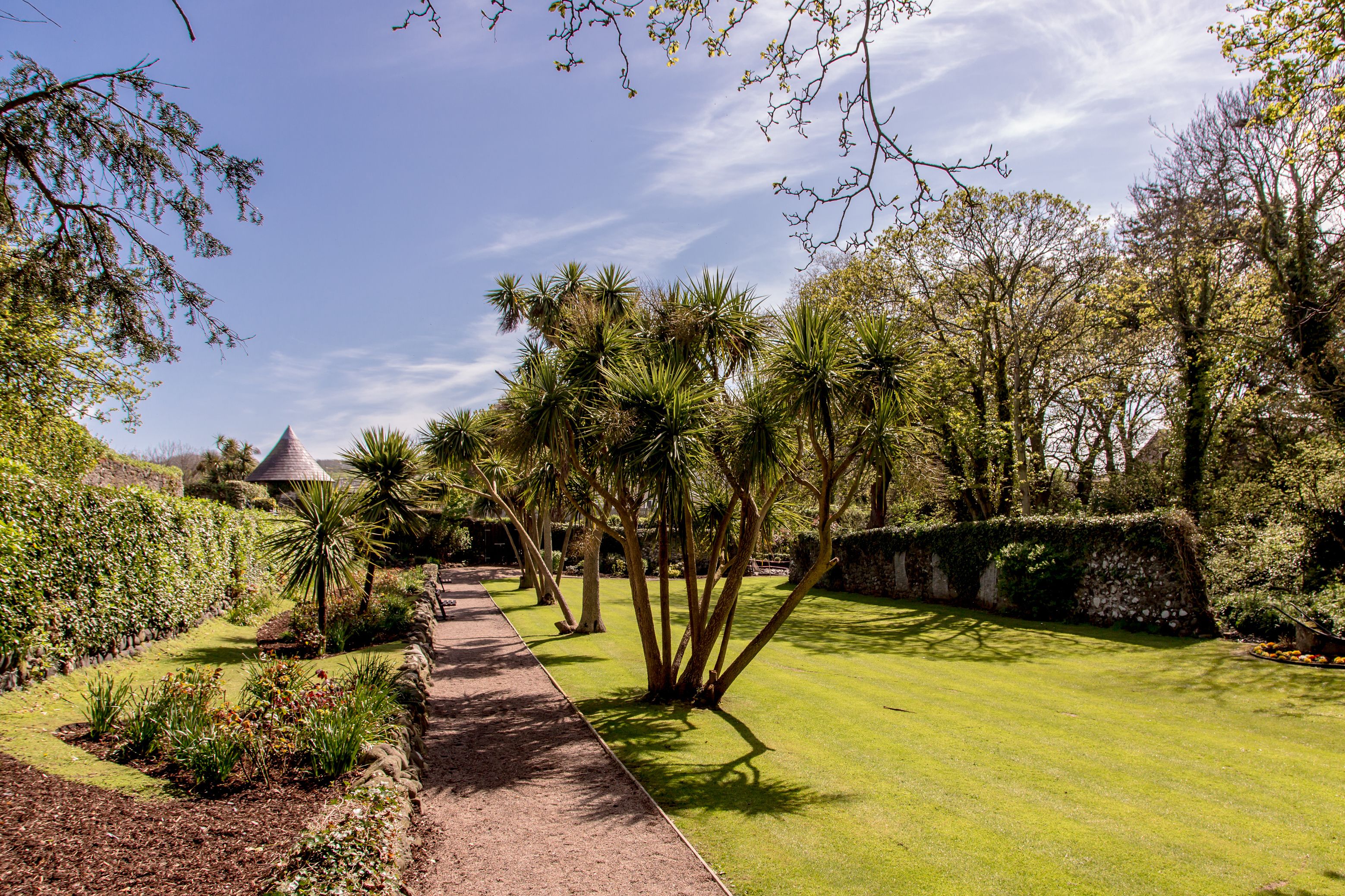 A garden pathway with a variety of plants and trees, a stone wall, and a small building with a conical roof.