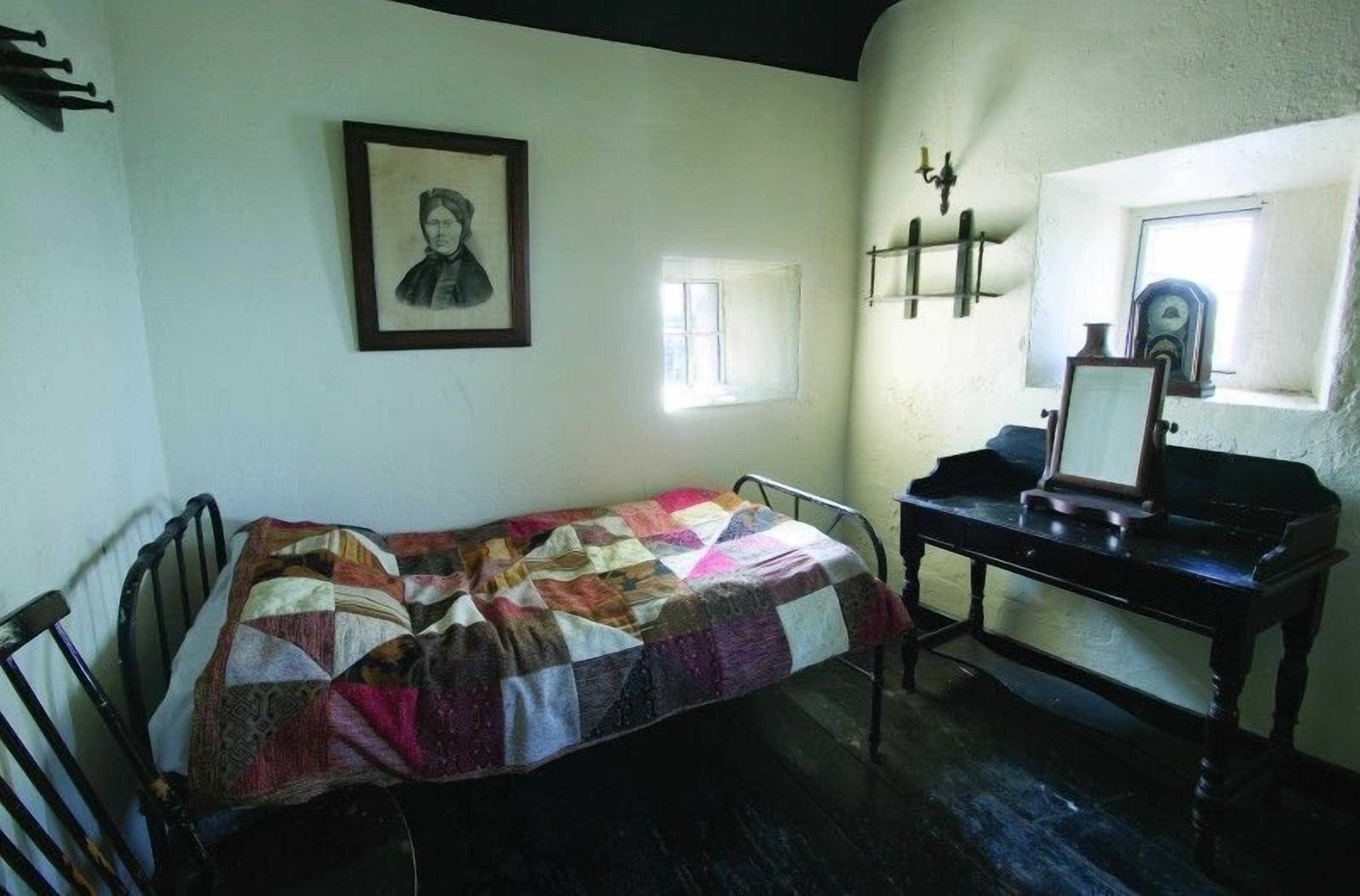 Una habitación con una cama, un escritorio, una silla, un cuadro en la pared, una ventana pequeña y un reloj.