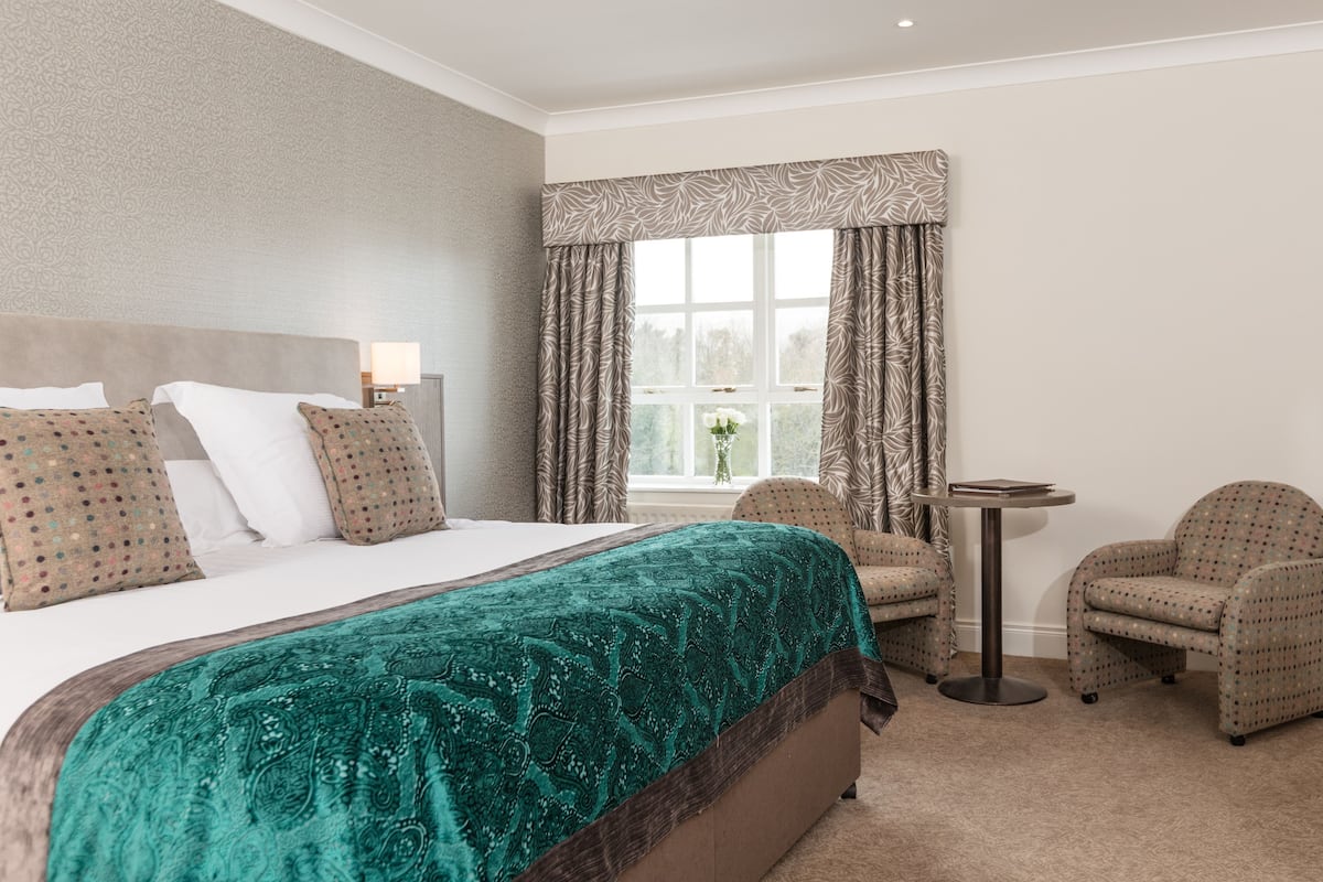 A hotel room with a large bed, two armchairs, a small table, and a window with curtains.