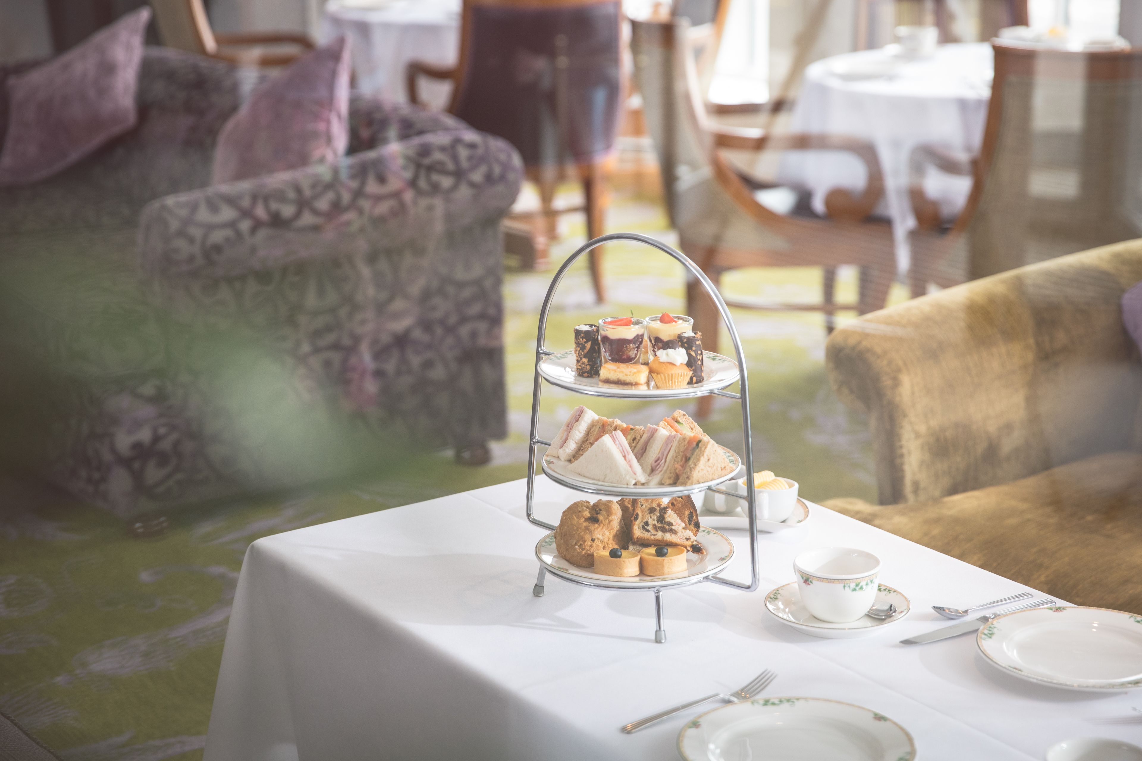 A three-tiered dessert stand with sandwiches and pastries on a table in a room with patterned sofas and a round table with a white tablecloth.