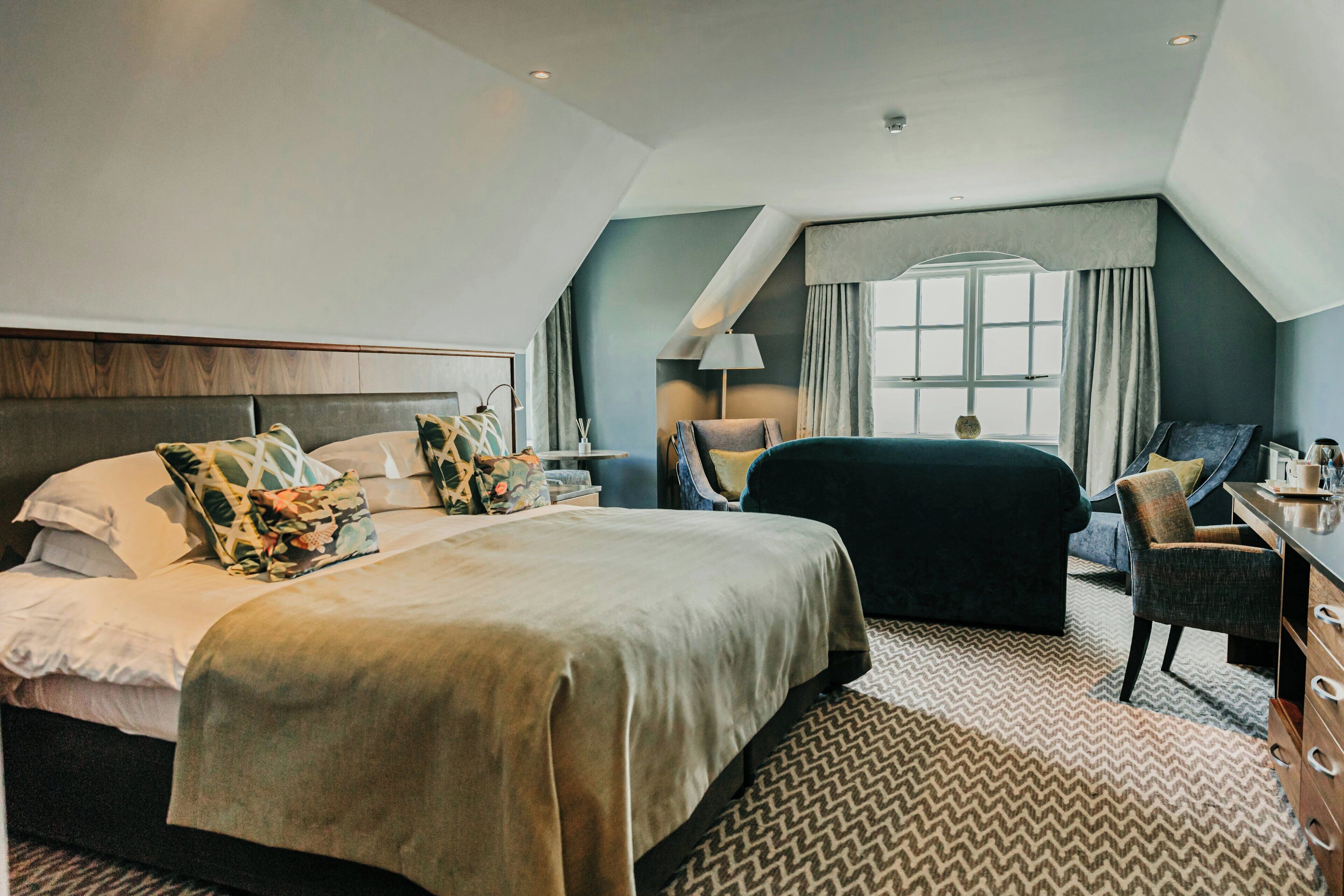 Habitación de hotel con una cama grande, un escritorio, una silla, un ventanal con cortinas y una alfombra estampada.