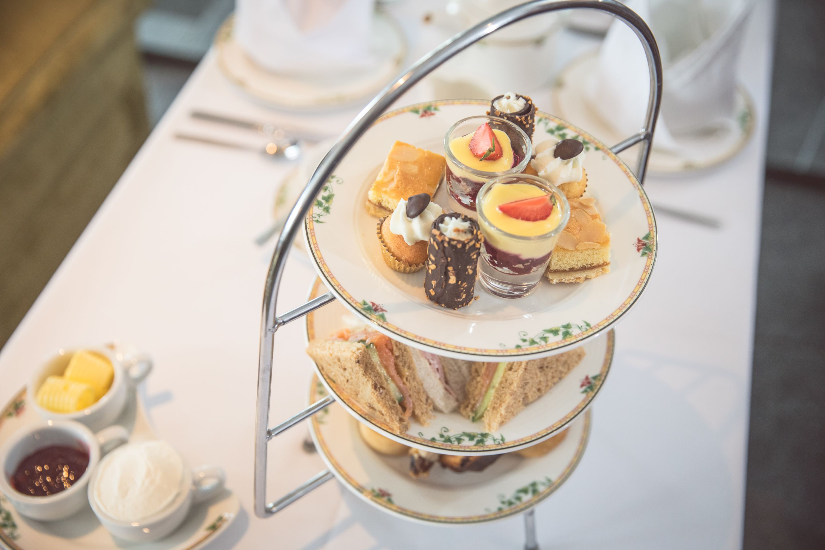 A three-tiered tea service stand with sandwiches, desserts, and beverages.