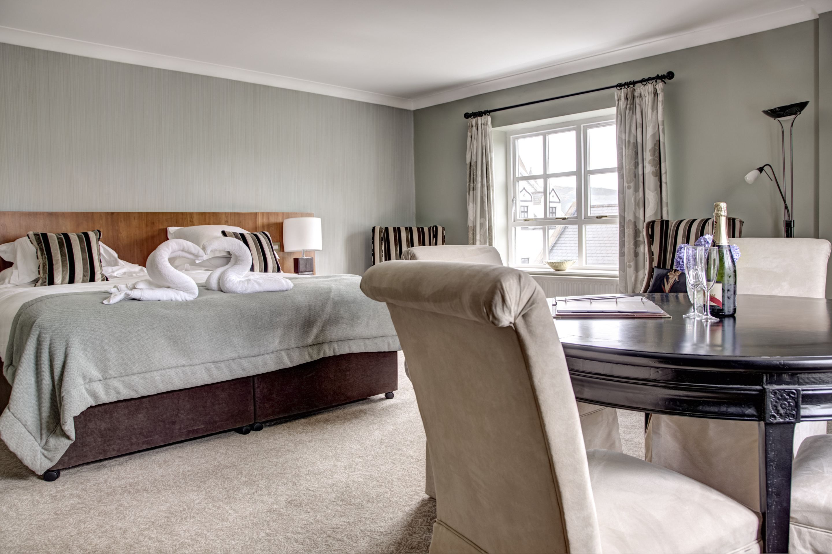 Una habitación de hotel con una cama, una mesa redonda con una silla, una ventana con cortinas y una botella de champán sobre la mesa.
