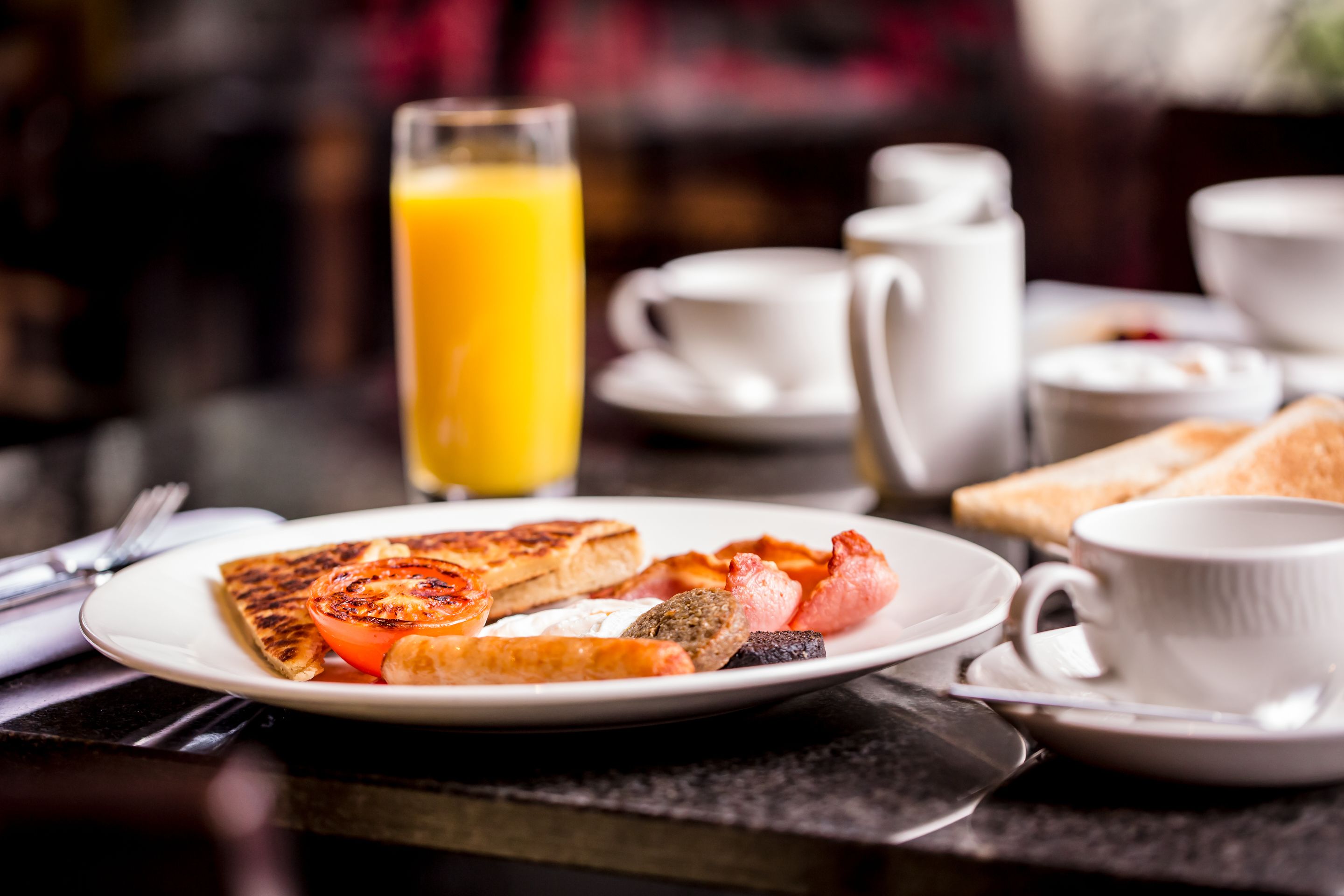 A breakfast setting with a plate of toast, eggs, and tomatoes, a glass of orange juice, and white cups and saucers on a table.