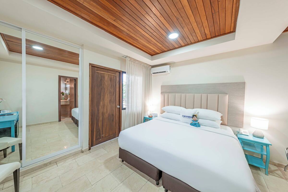 A modern hotel room with a large bed, wooden ceiling, and a sliding glass door leading to a bathroom.