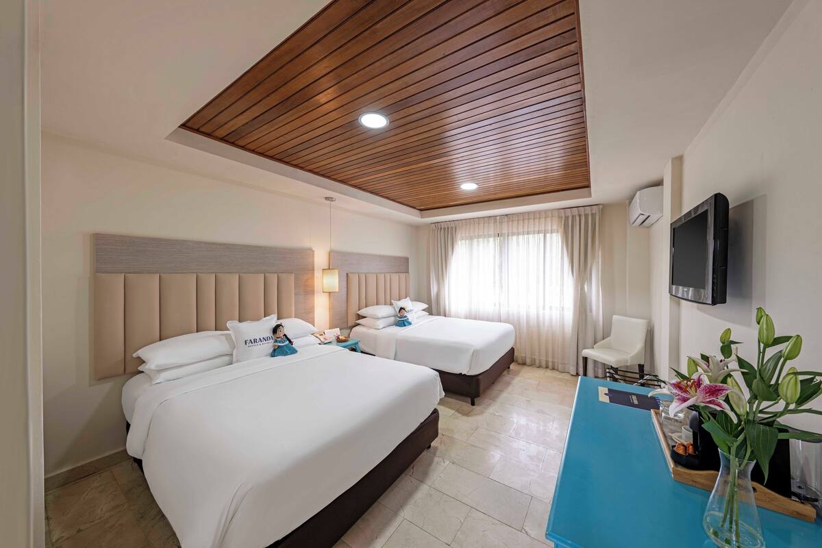 A hotel room with two beds, a wooden ceiling, a TV, and a desk with a vase of flowers.