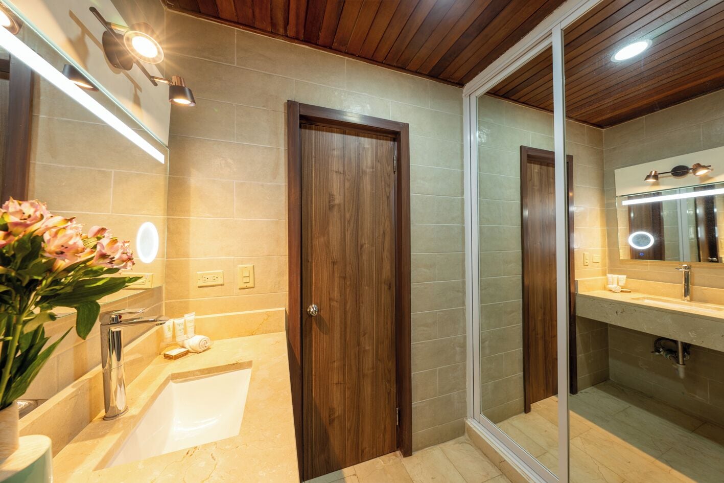 A modern bathroom with a wooden door, a glass shower enclosure, a sink with toiletries, and a mirror with lights.