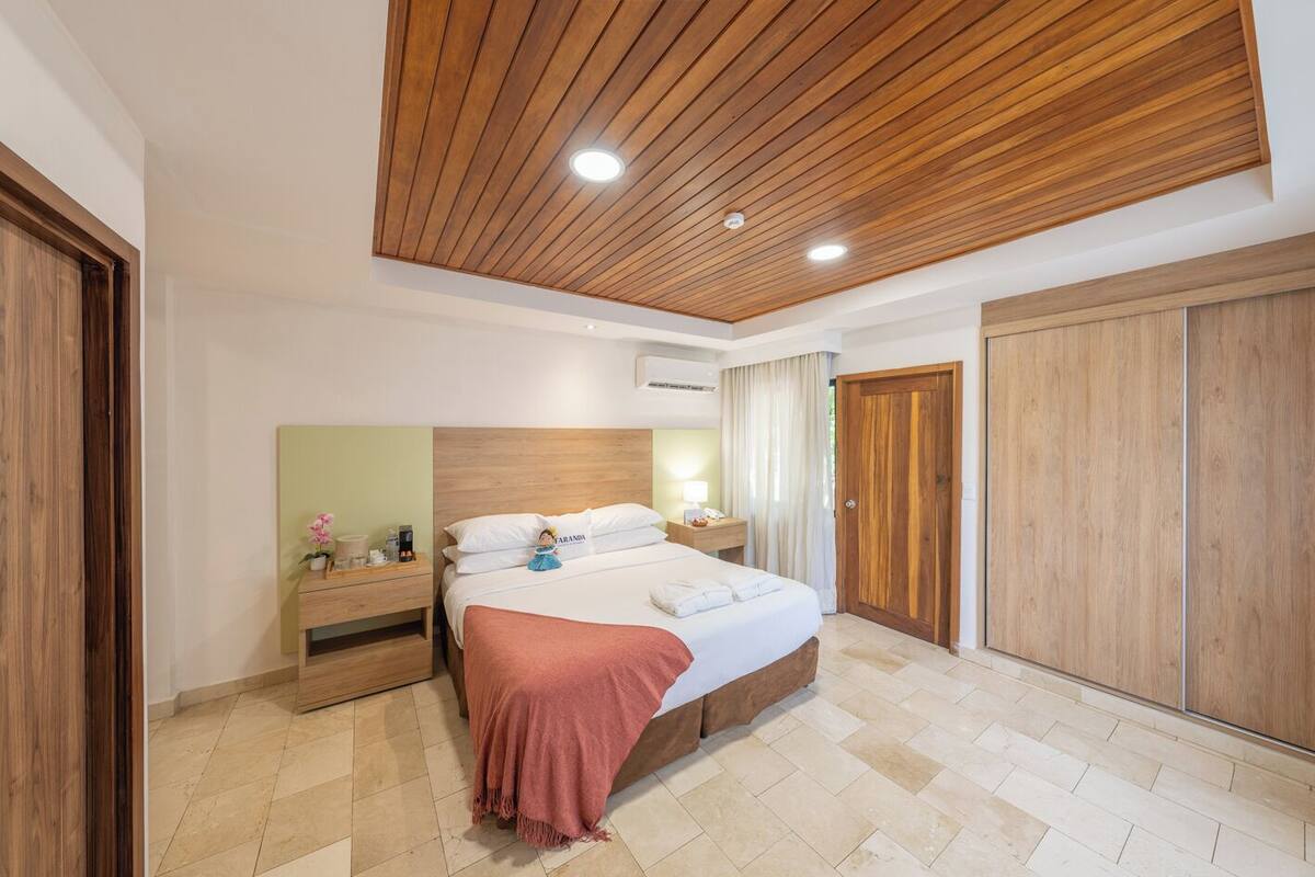 A bedroom with a wooden ceiling, a bed with a pink blanket, a nightstand with a lamp, and a wardrobe.
