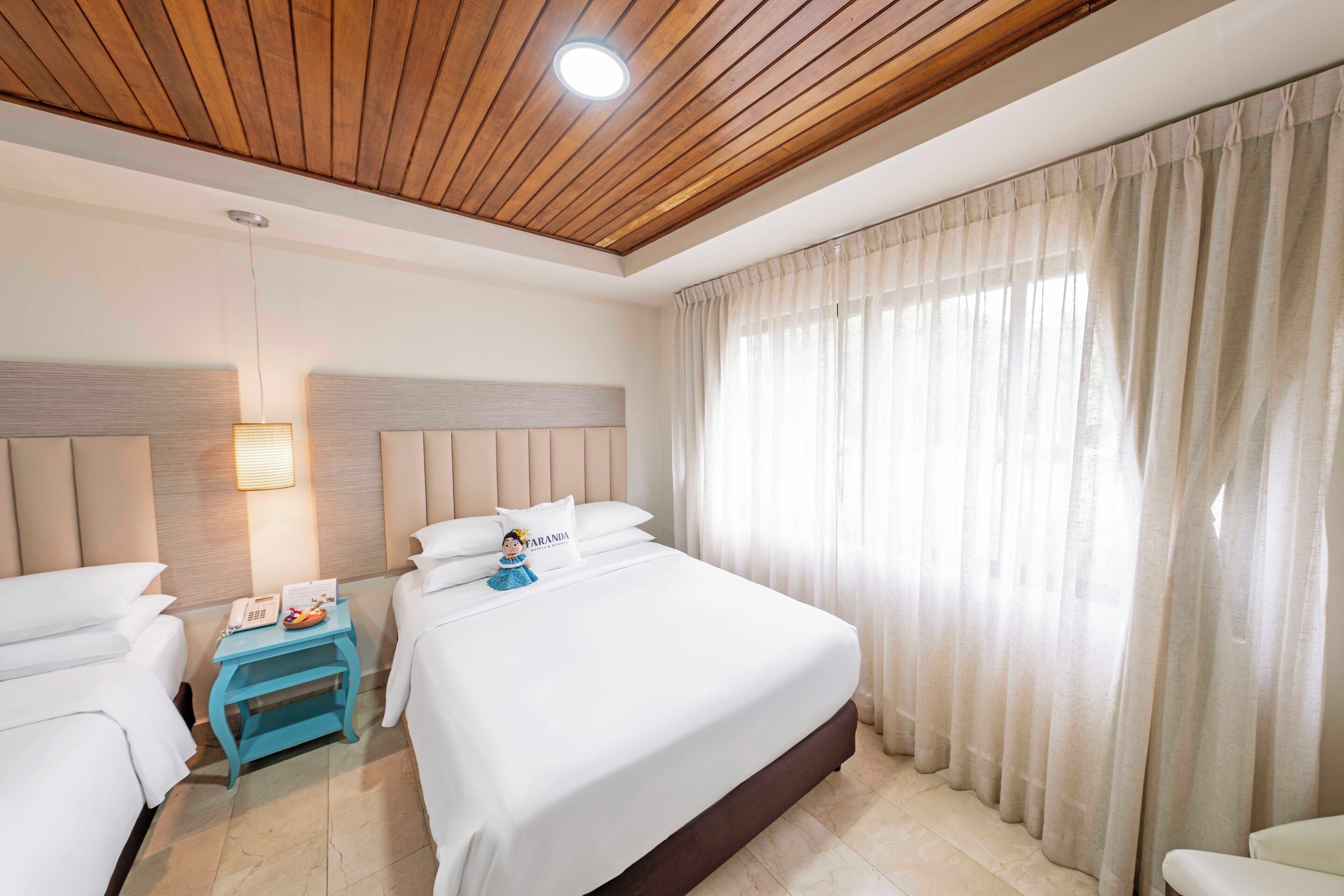 A hotel room with a large bed, two bedside tables, a nightstand, a wall-mounted lamp, and a window with curtains.