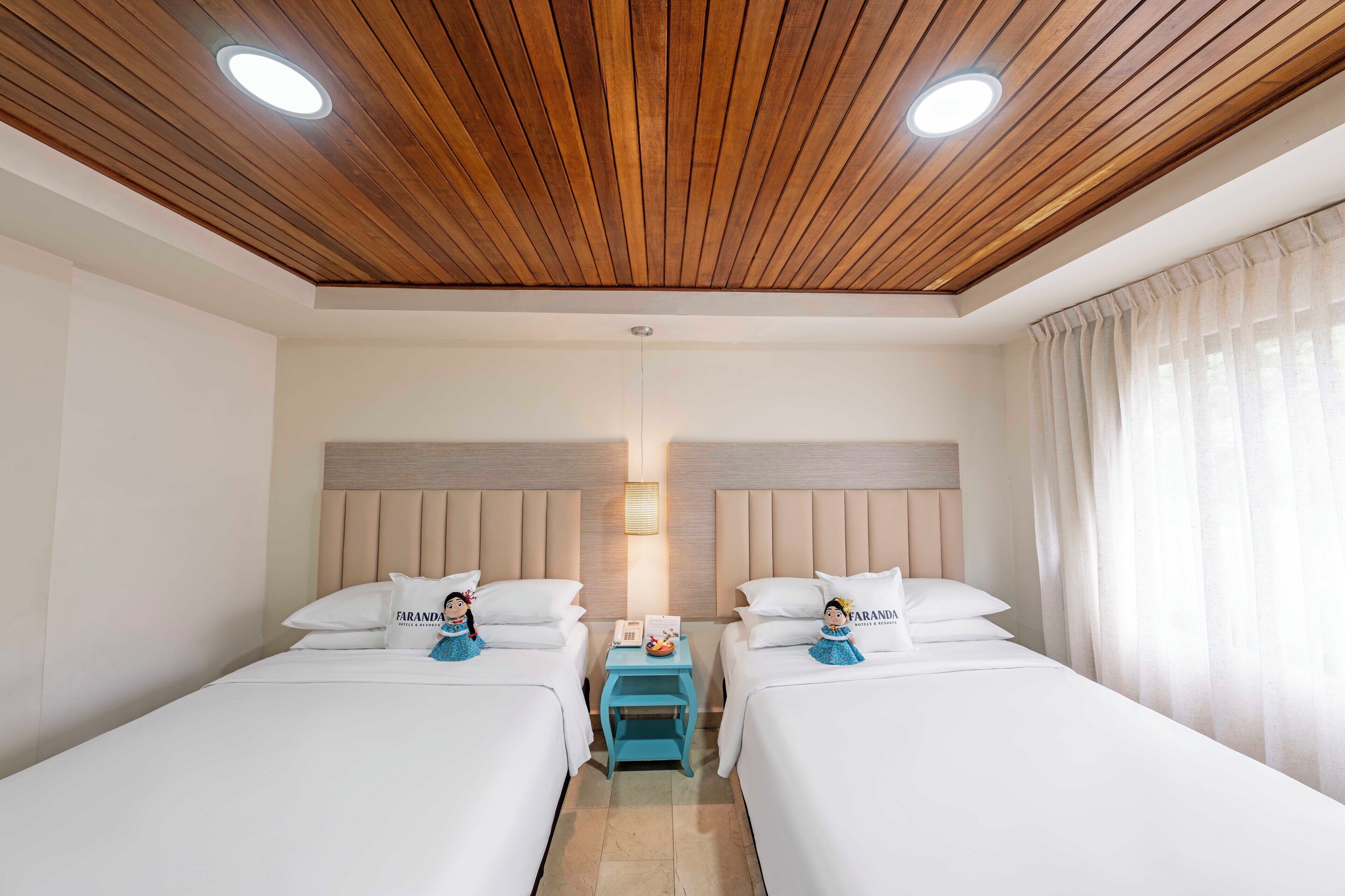 A hotel room with two beds, wooden ceiling, a small table with a lamp, and a window with curtains.