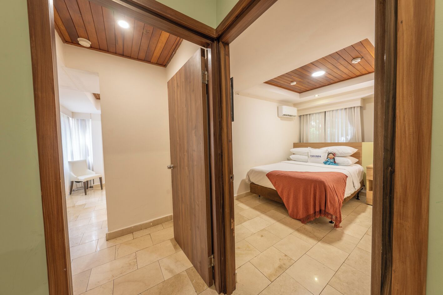 A bedroom with a wooden ceiling, a bed with a pink blanket, and a white chair in the adjacent room.