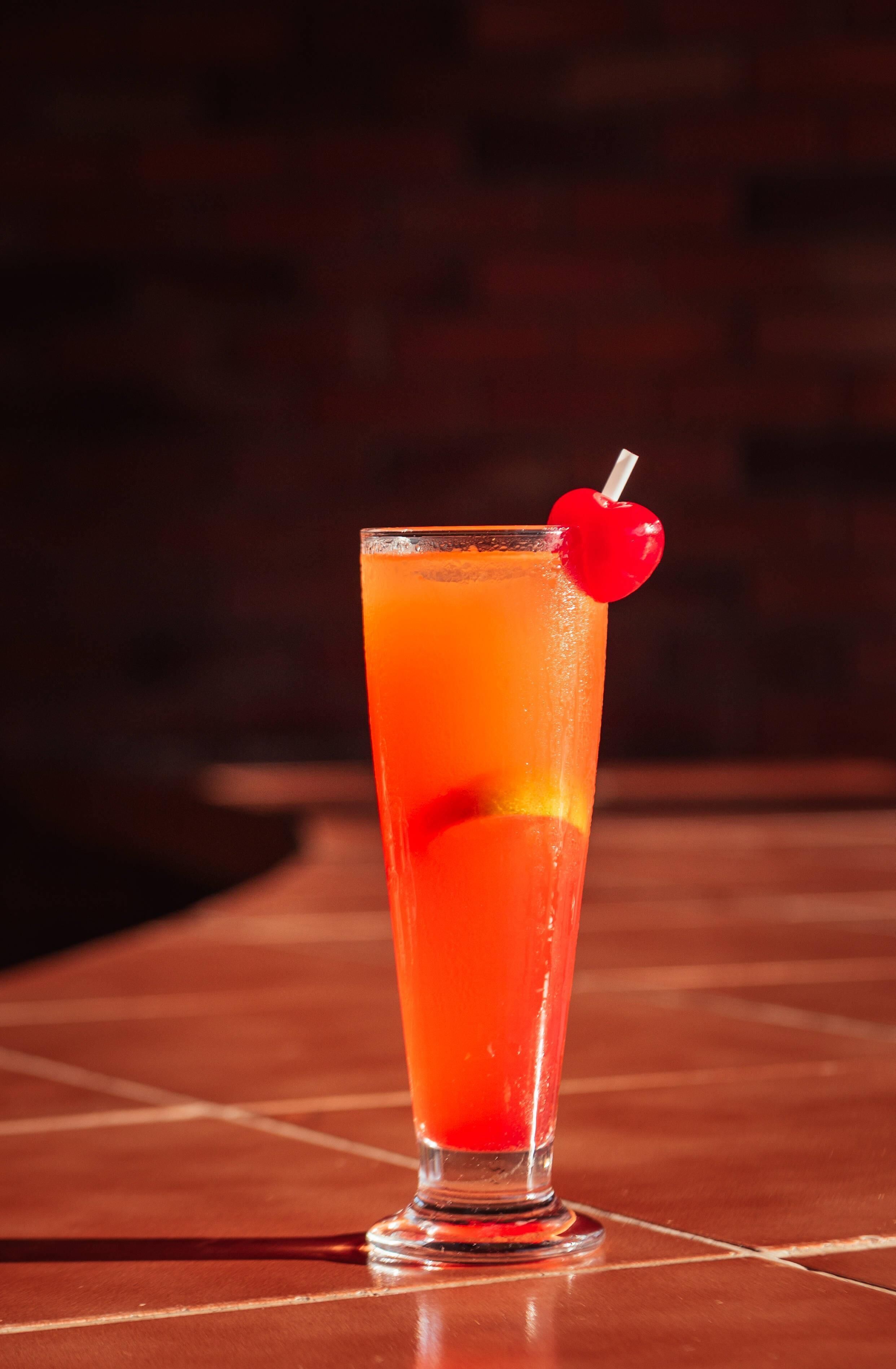 Un vaso alto con una bebida roja y una cereza en un palito.