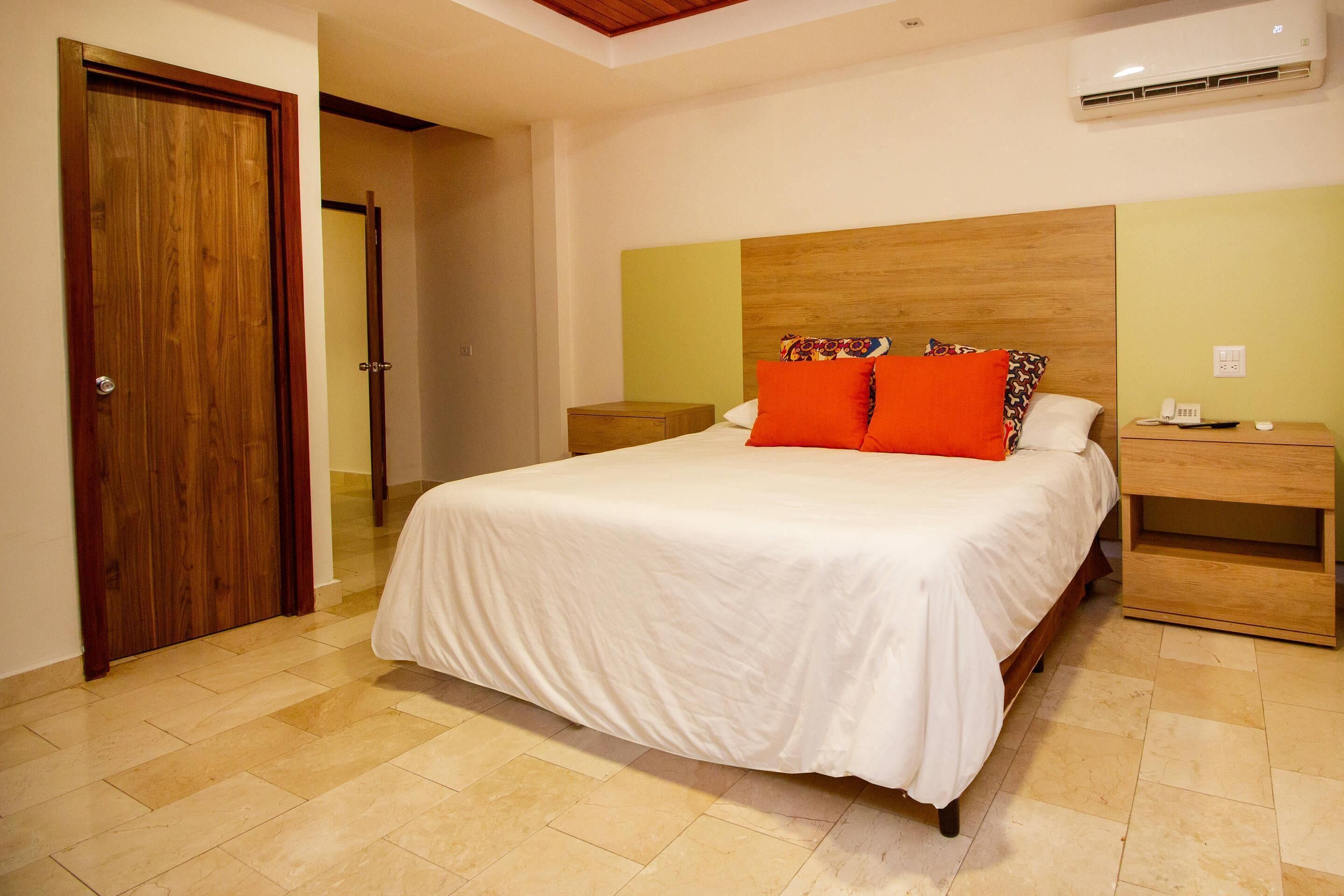 Un dormitorio con cabecera de madera, cubrecama blanco, dos mesitas de noche y una mesita auxiliar de madera.