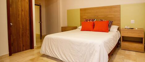 Un dormitorio con cabecera de madera, cubrecama blanco, dos mesitas de noche y una mesita auxiliar de madera.