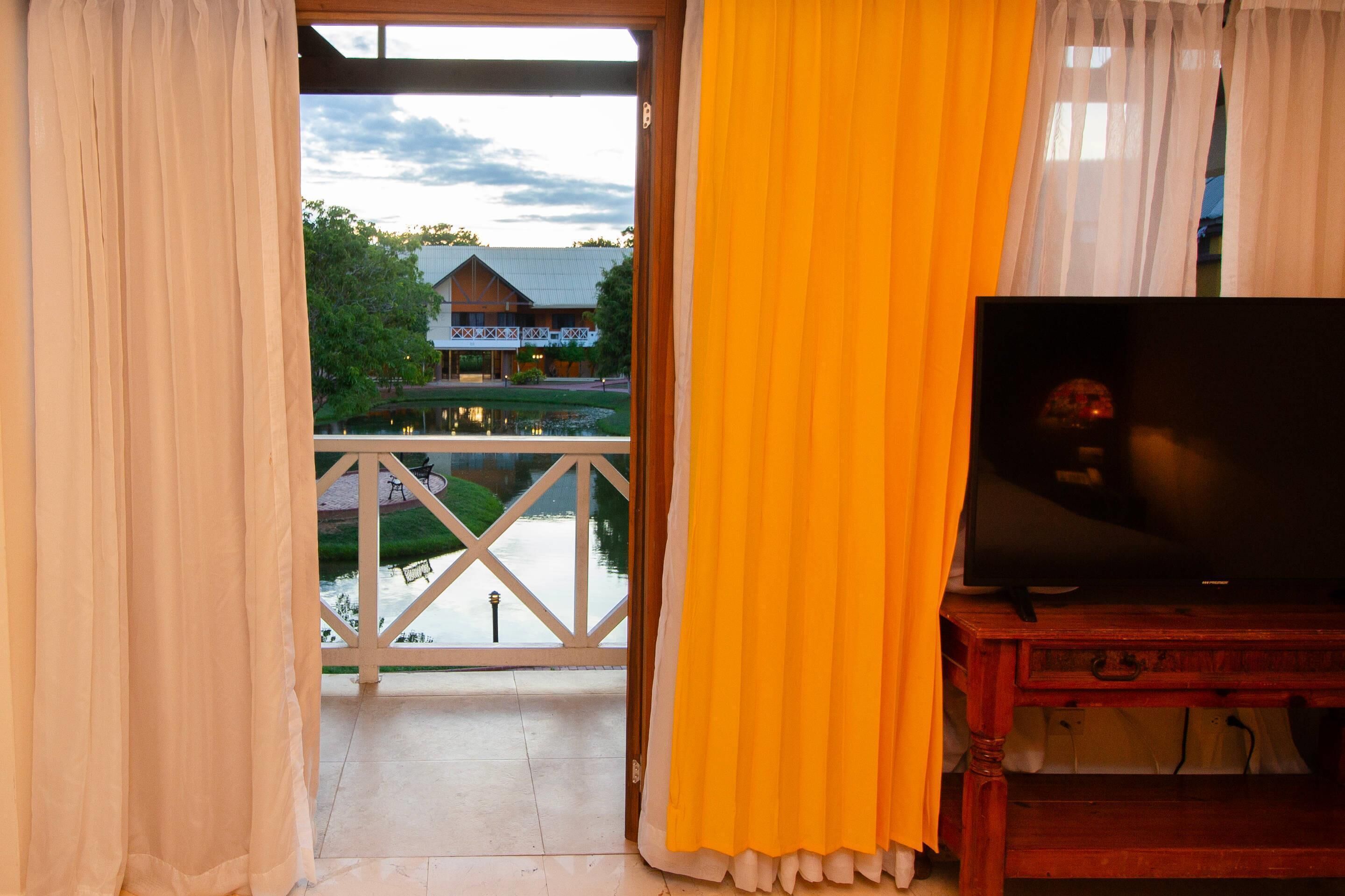 Ein Zimmer mit Fernseher auf einem Holzständer, ein Balkon mit Geländer und Blick auf ein Gebäude und Bäume.