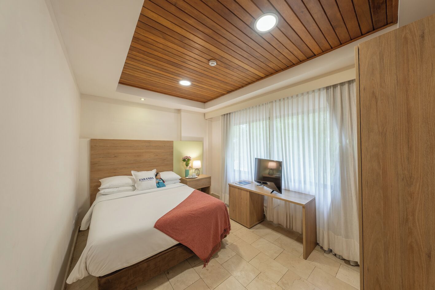 A hotel room with a wooden ceiling, a bed with white bedding, a desk with a TV, and a window with curtains.