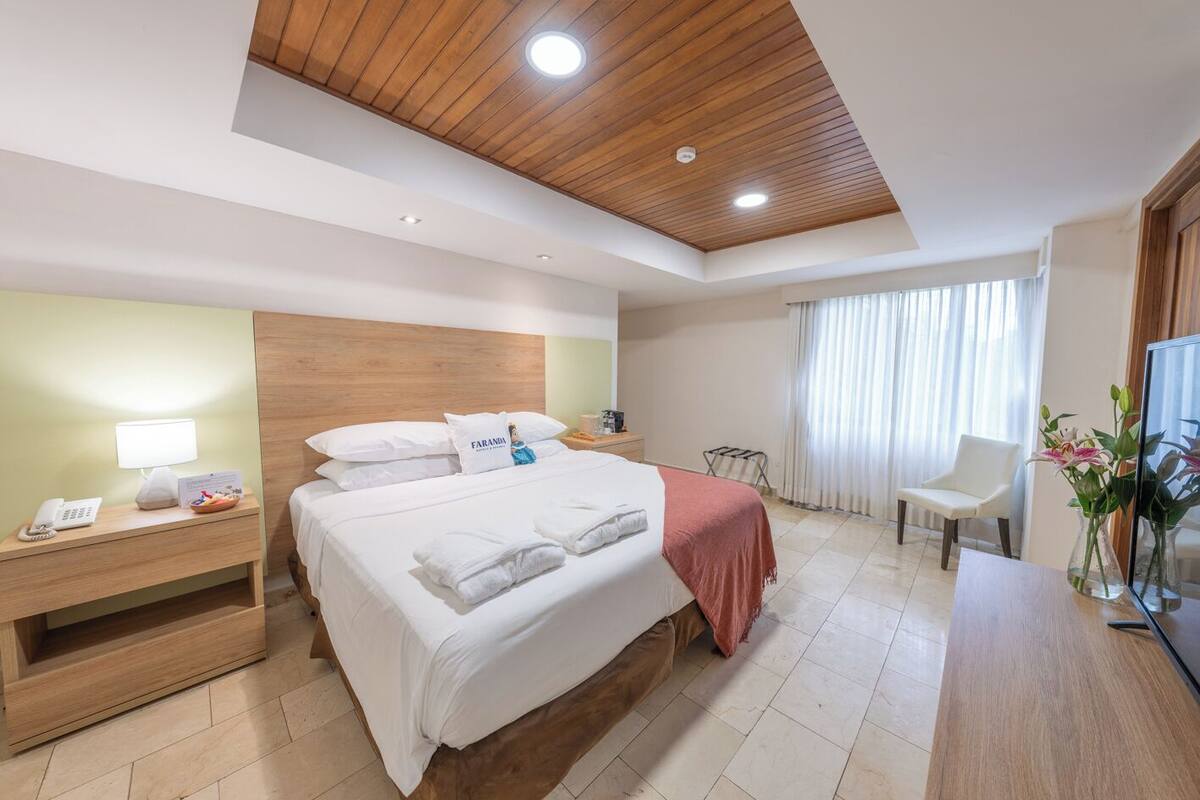 A hotel room with a wooden ceiling, a bed with white linens, a nightstand with a lamp, a desk with a chair, and a window with curtains.