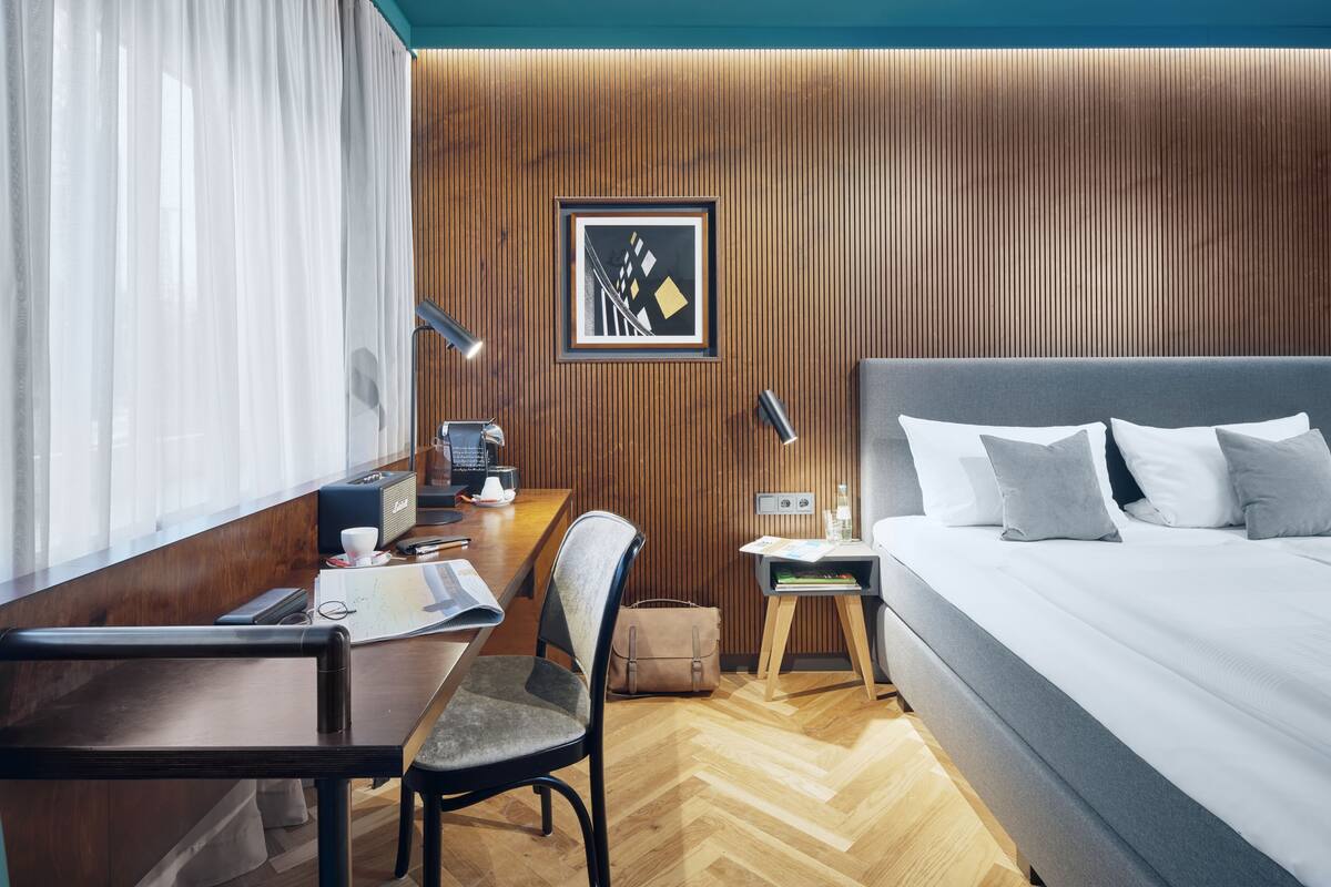 A hotel room with a wooden panel wall, a bed with grey bedding, a desk with a chair, a nightstand, and a framed picture on the wall.