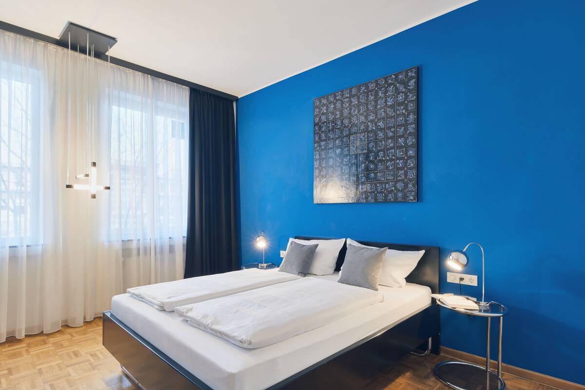 A modern hotel room with a large bed, a bedside table with a lamp, and a blue accent wall.