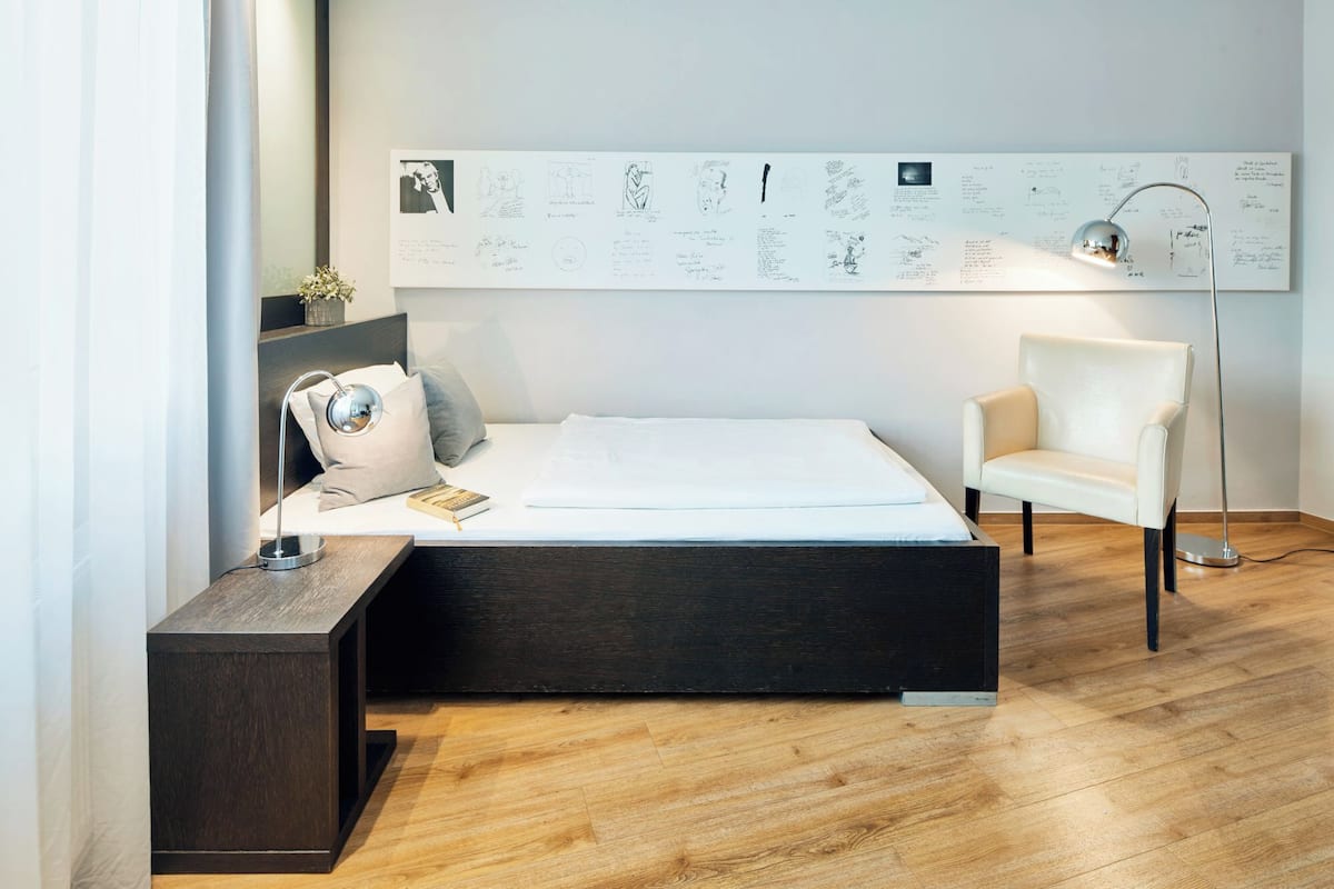 A modern bedroom with a large bed, a side table, a chair, and a floor lamp. There is a wall-mounted artwork above the bed.