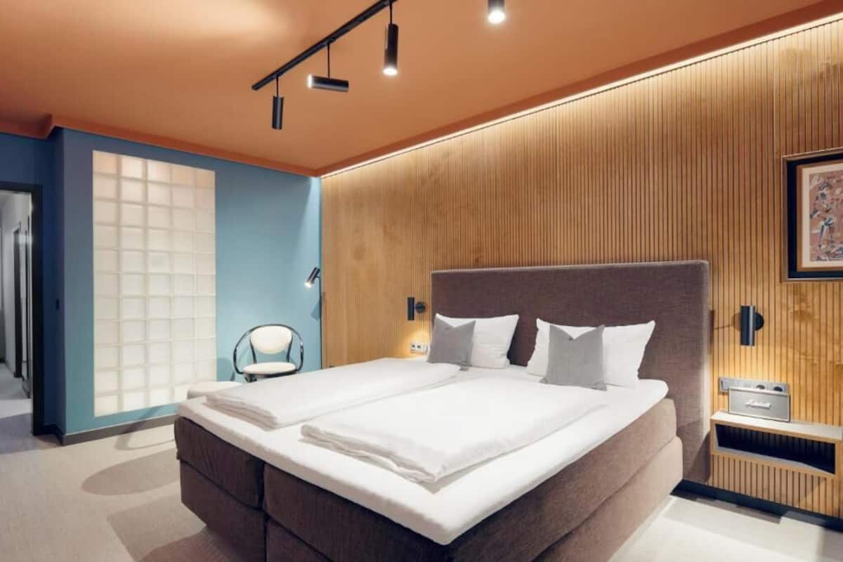 A modern hotel room with a large bed, a bedside table, a chair, and a wall-mounted TV.