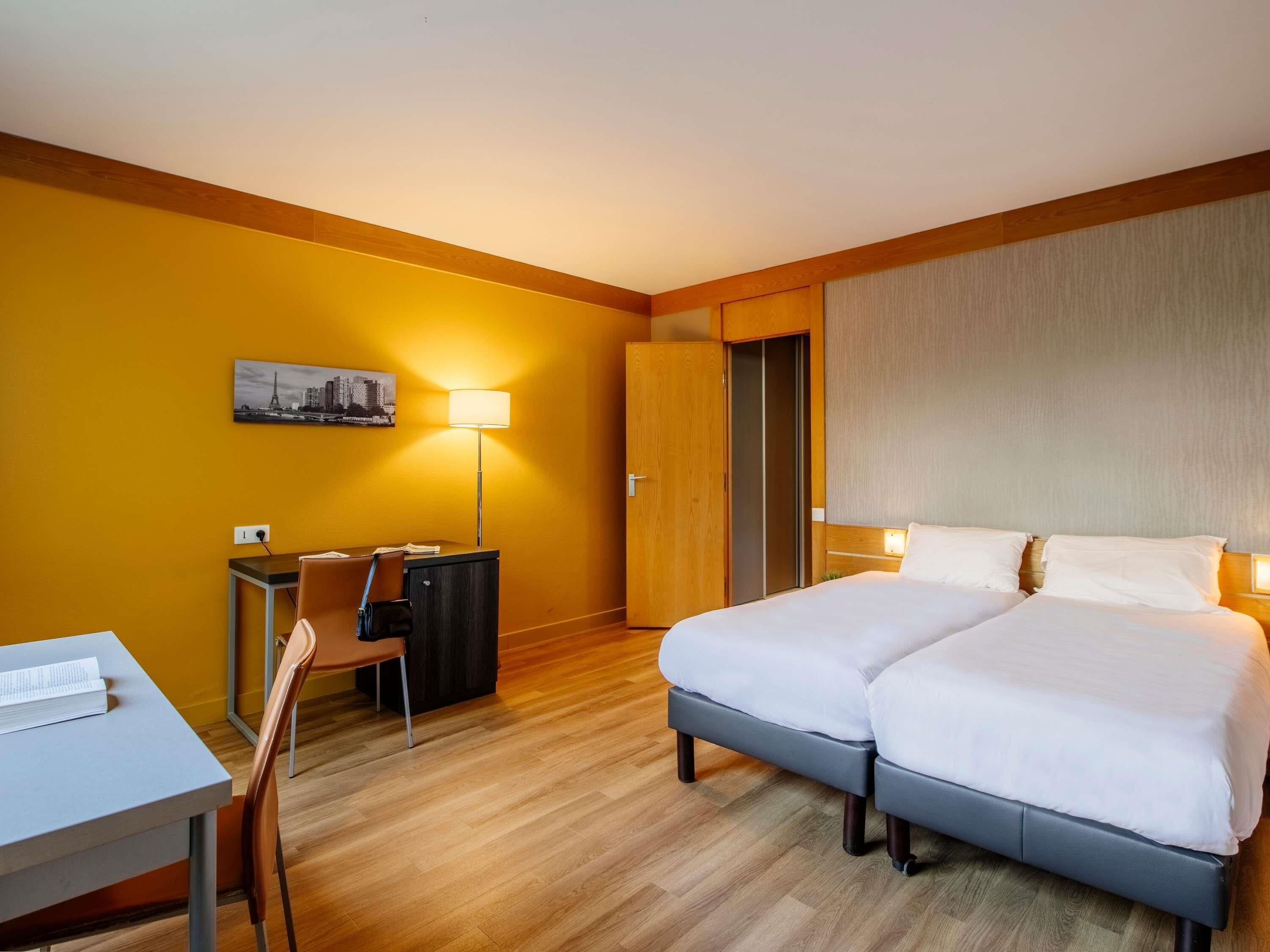 Quarto de hotel com cama, escrivaninha, cadeira, luminária, guarda-roupa e uma pintura com vista para a cidade.