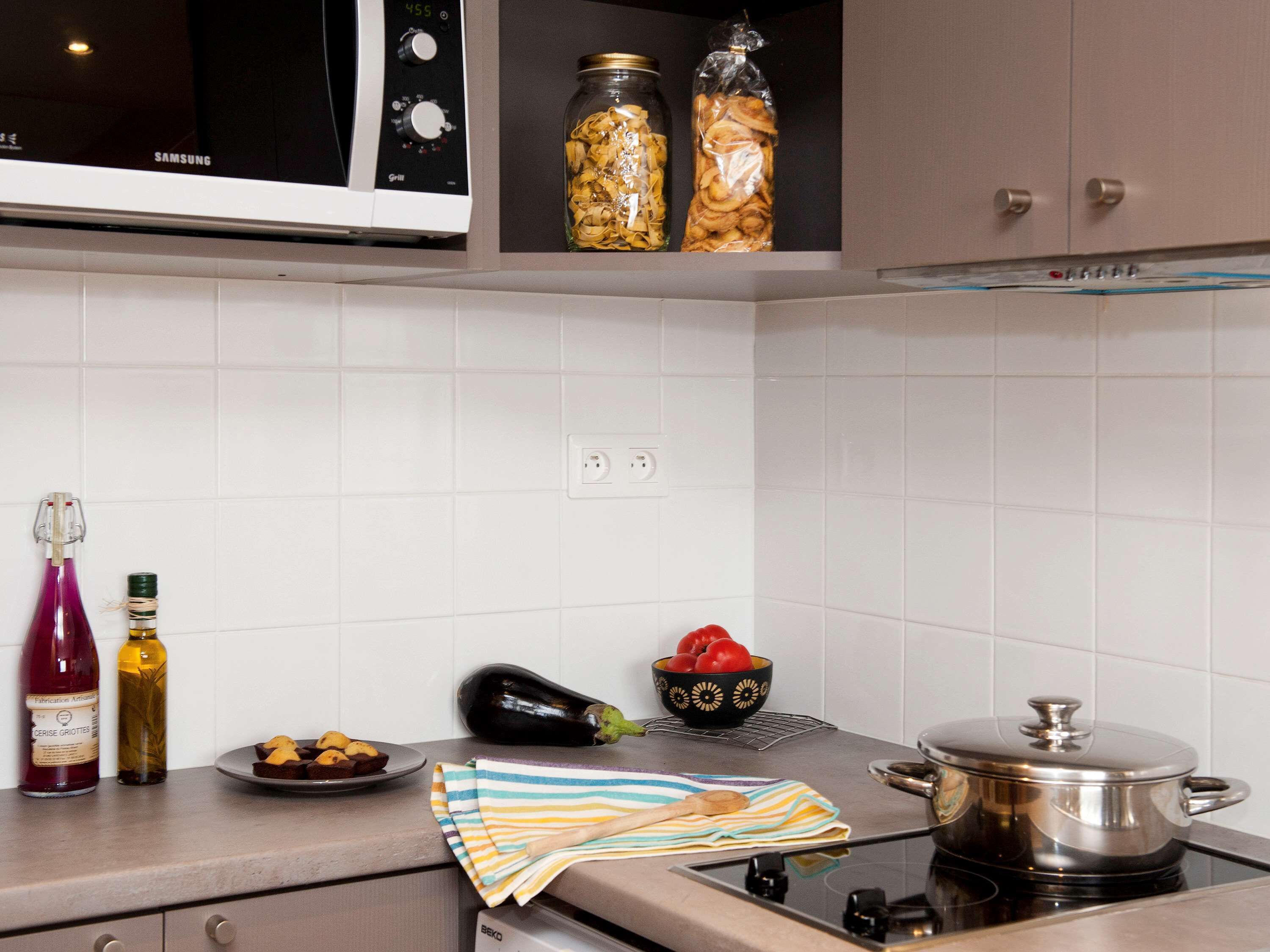Une cuisine équipée d’un four à micro-ondes, de bocaux contenant des aliments, d’une bouteille de vin, d’un saladier rempli de tomates, d’une aubergine et d’une casserole sur la cuisinière.