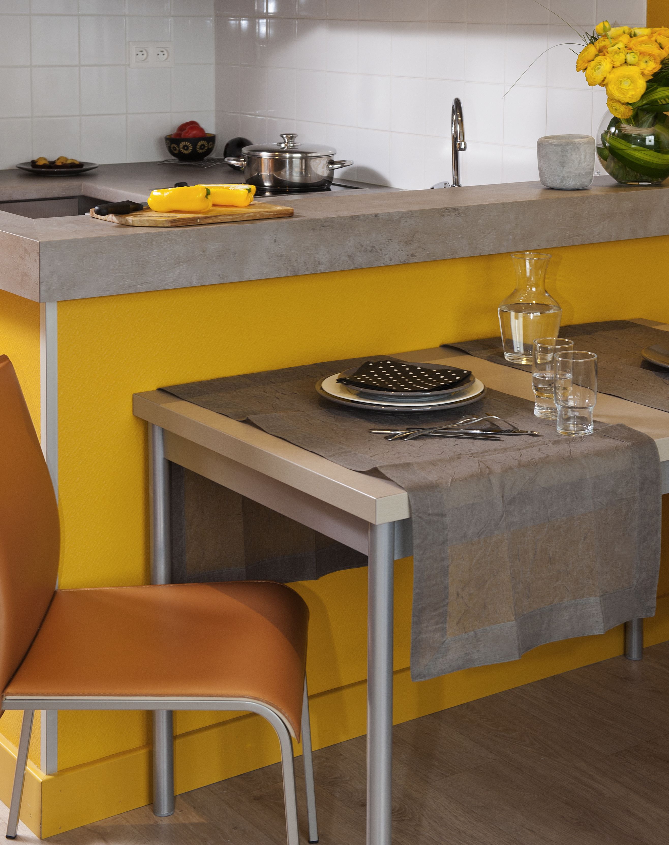 Cucina con parete gialla, piano di lavoro grigio, lavello, fornello e zona pranzo con tavolo e sedia.