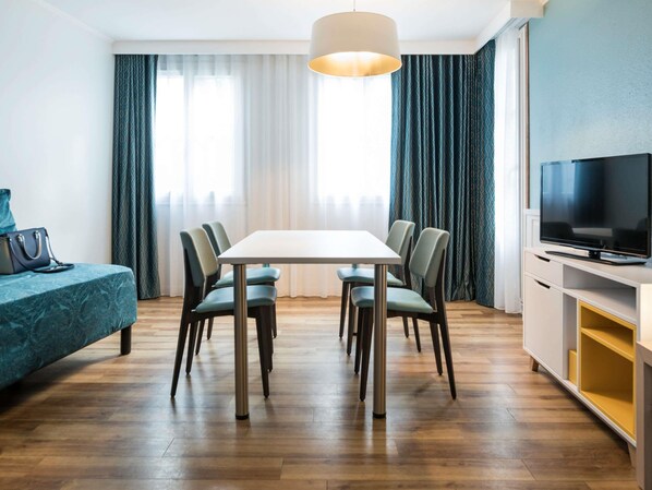 Una habitación de hotel moderna con una mesa de comedor, sillas, un sofá, un televisor y un ventanal con cortinas.