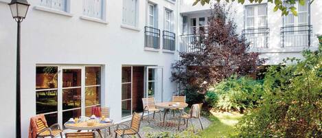 A courtyard with a table and chairs, a lamp post, and a building with large windows and shutters.