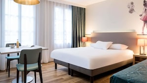 Quarto de hotel com cama, secretária, cadeira, luminária e janela com cortinas.