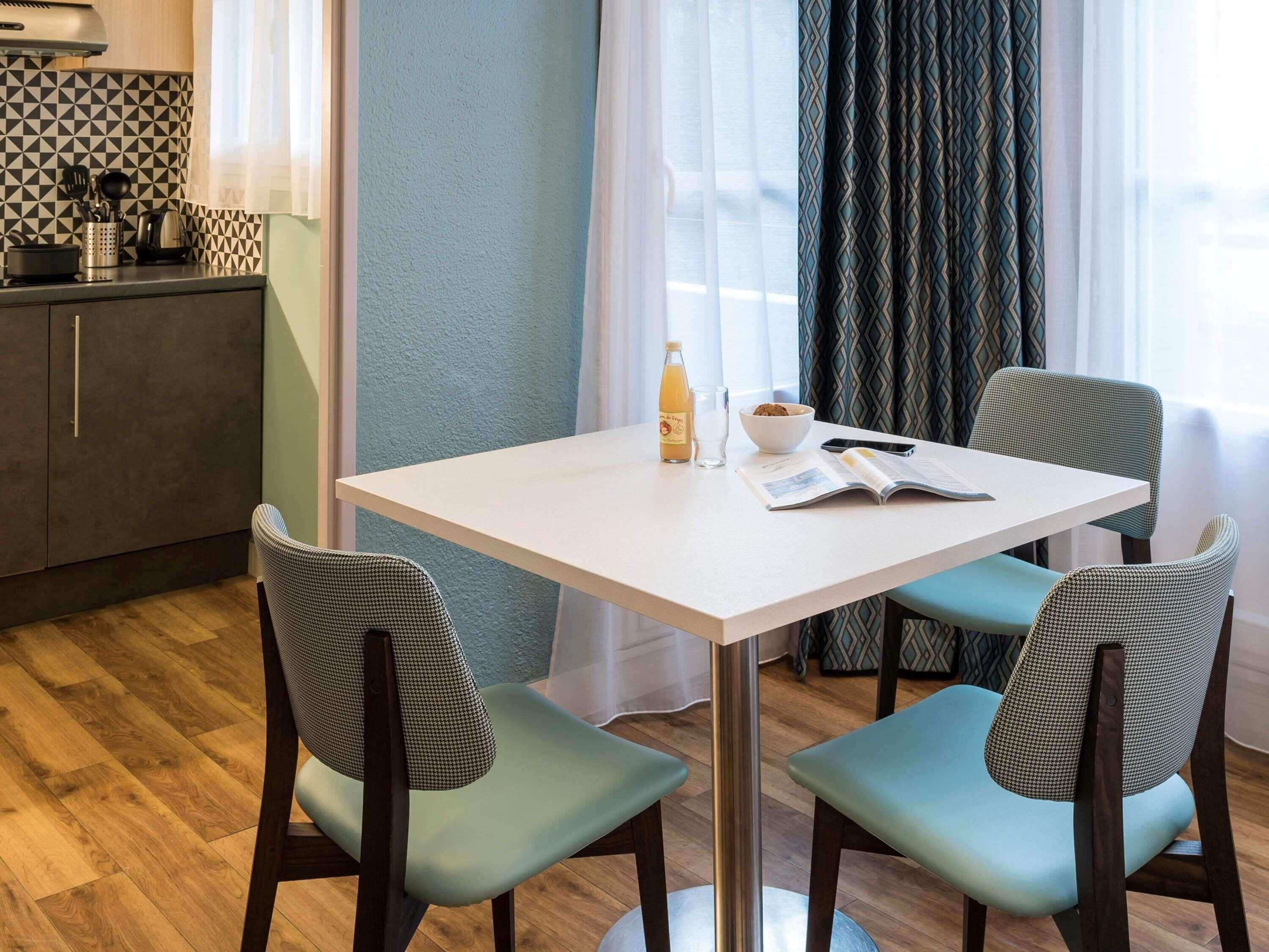 A small dining area with a white table, two patterned chairs, a bowl of cookies, a bottle of orange juice, and a book.