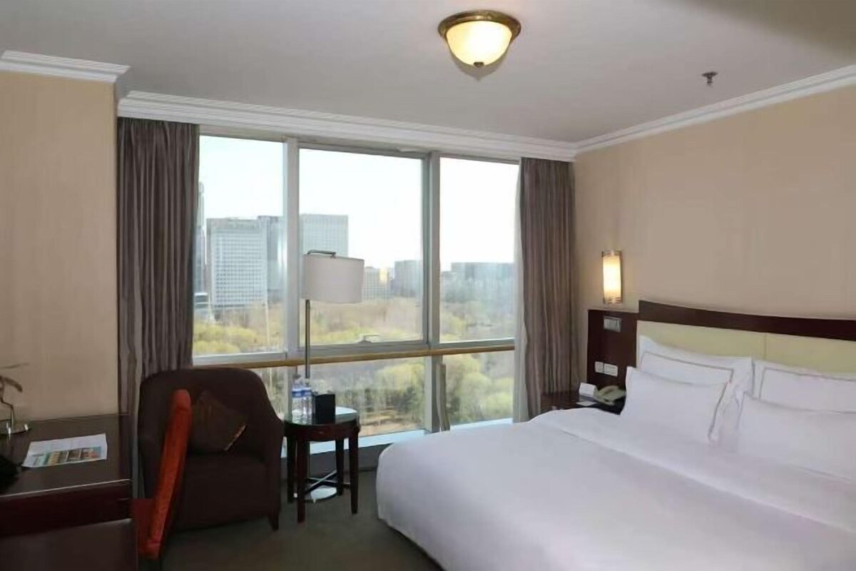 A hotel room with a large bed, a desk with a chair, a lamp, and a view of the city through large windows.