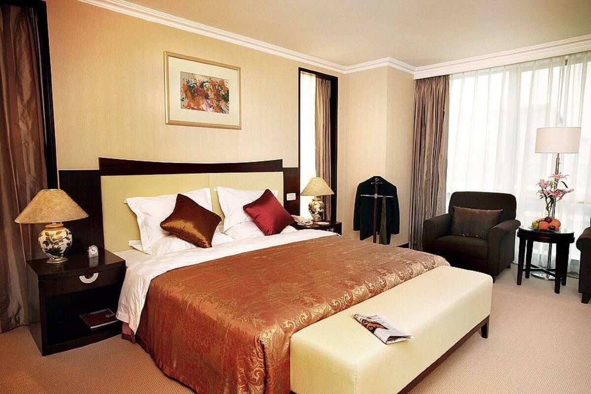 Una habitación de hotel con una cama grande, una mesita de noche con lámpara, una silla, una pequeña mesa con lámpara y una ventana con cortinas.