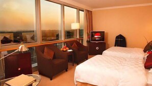 A hotel room with a large bed, a desk with a lamp, a TV, and a view of the city through large windows. - Beijing Jinjiang Fuyuan Hotel (Beijing)