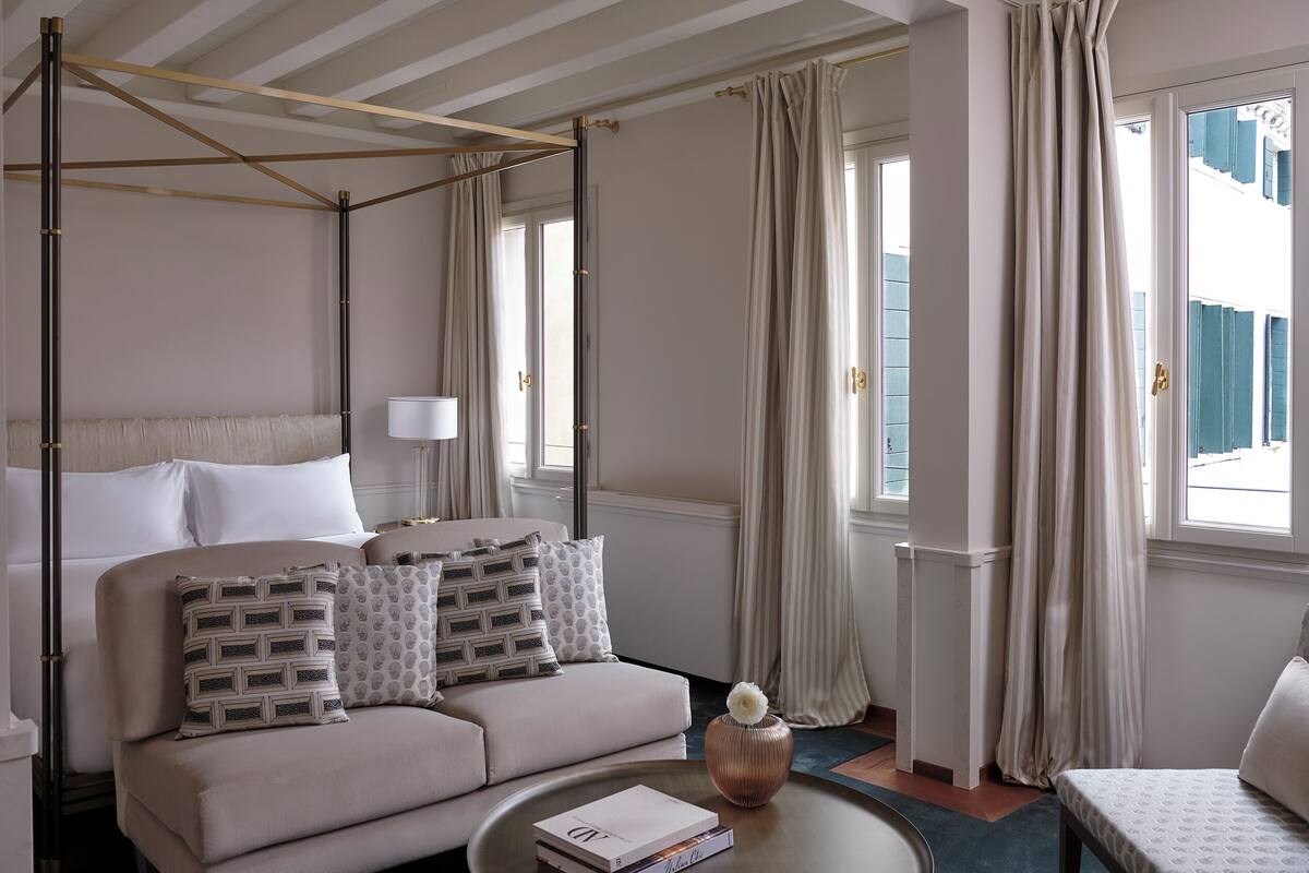A hotel room with a four-poster bed, a sofa, a coffee table with books, and a view of the outside through large windows.