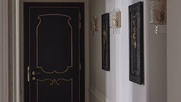 A hallway with a black door featuring gold trim, a white wall with framed artwork, and decorative wall sconces.