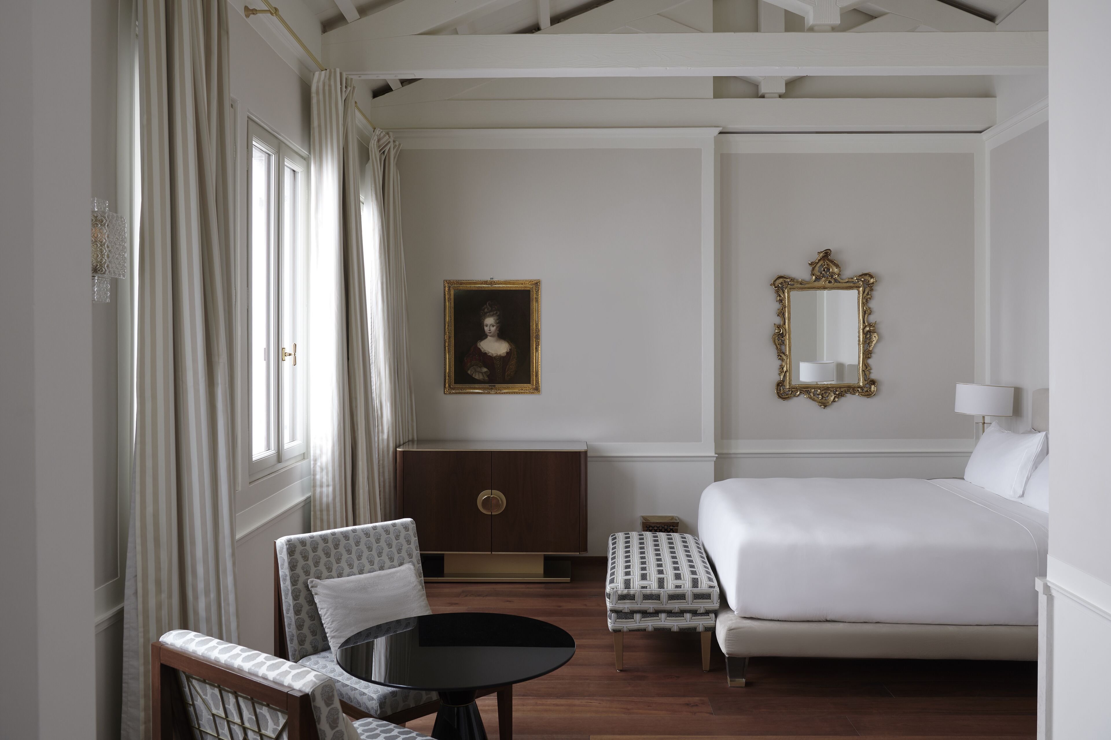 Une chambre d’hôtel avec un lit, une chaise, une petite table, une armoire, un tableau et un miroir.